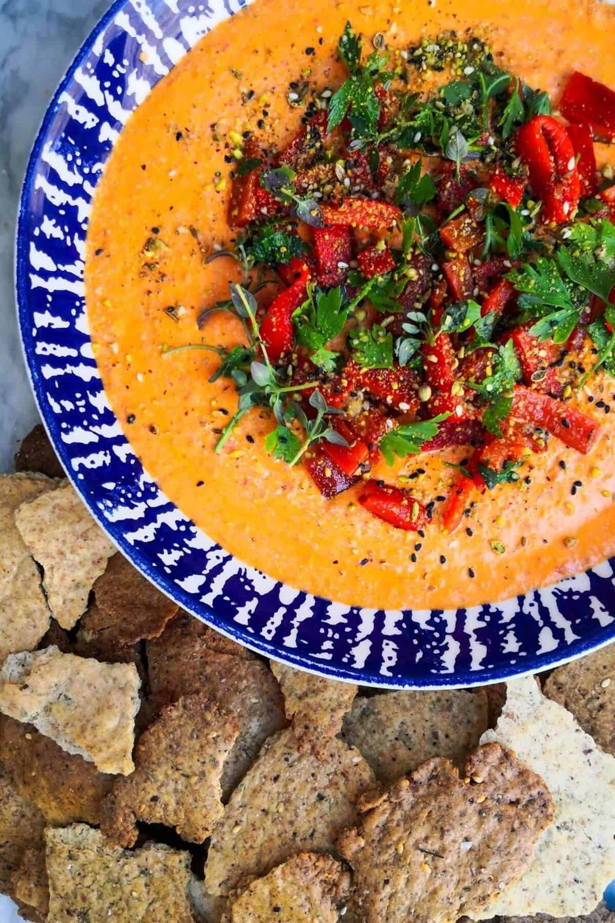 Red pepper and feta dip in a bowl with blue and white stripes, topped with red pepper, parsley and dukkah with crackers on the side