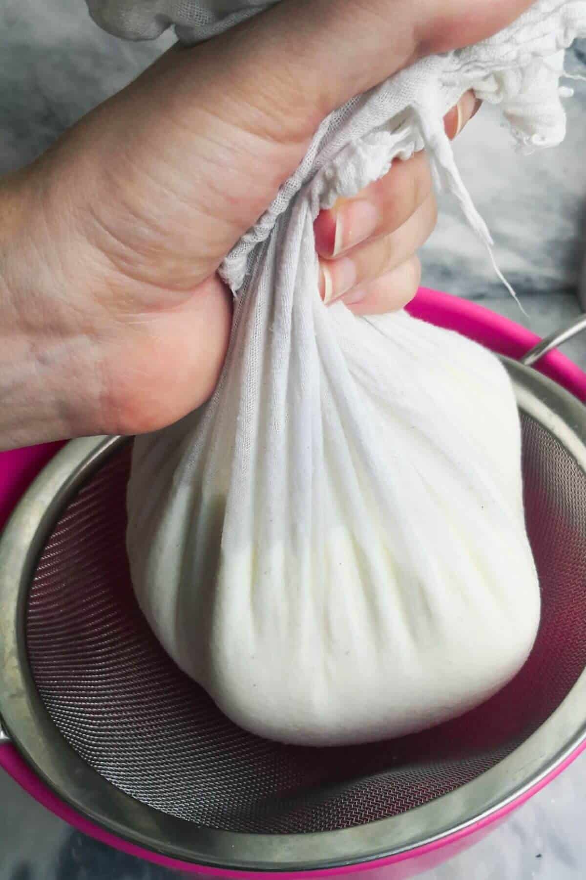 Hand bundling up cheesecloth with yogurt in it over a silver sieve.