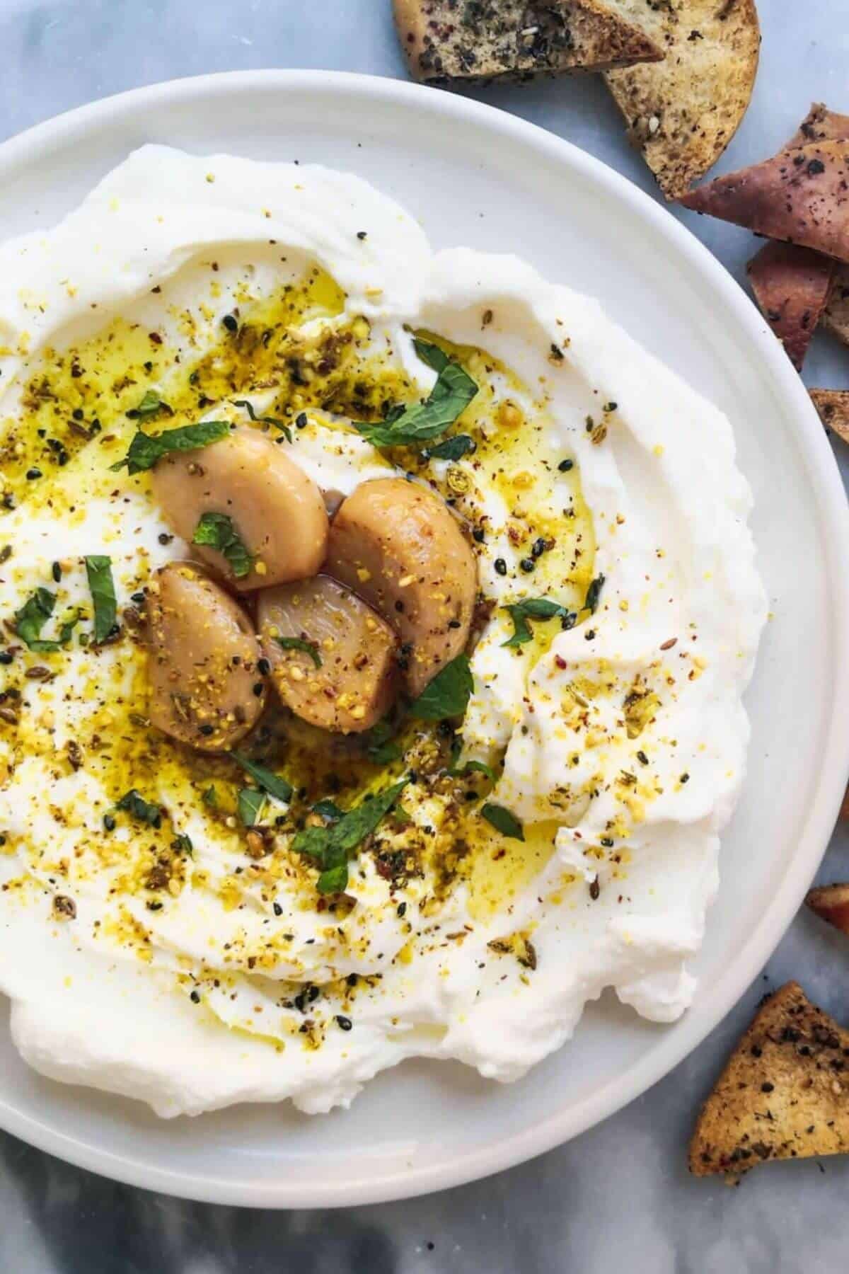 Labneh with confit garlic and dukkah on a plate with pita chips on the side.