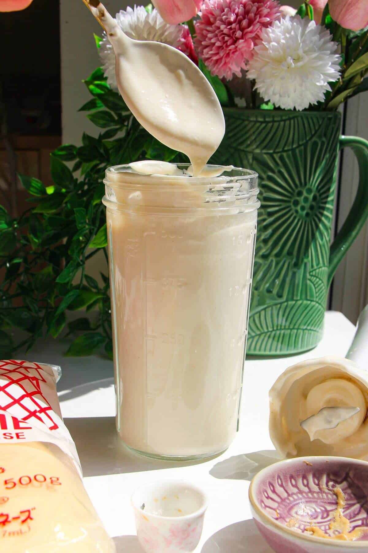 Kewpie mayo in a tall glass jar on a wooden board with a spoon on top.