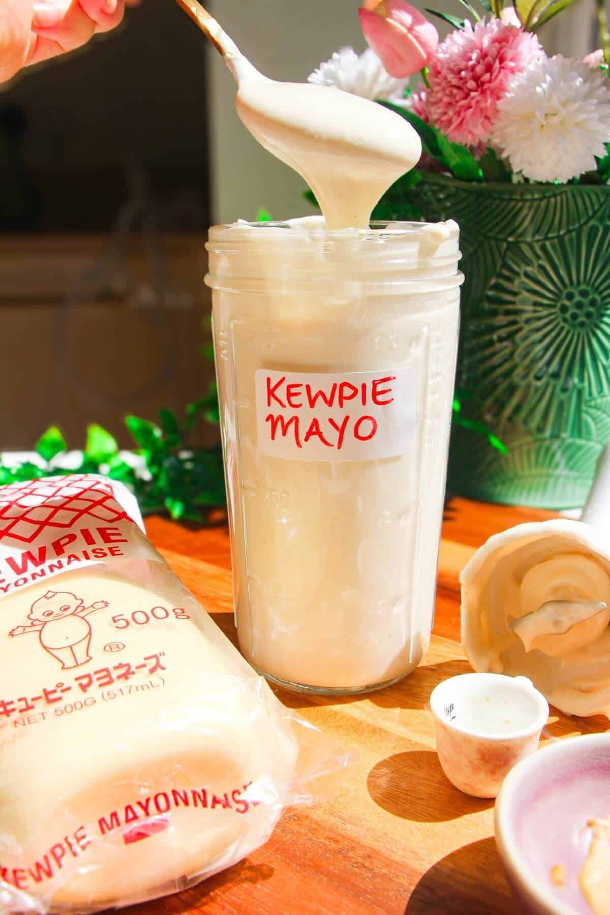 Kewpie mayo in a tall glass jar on a wooden board with a spoon on top.