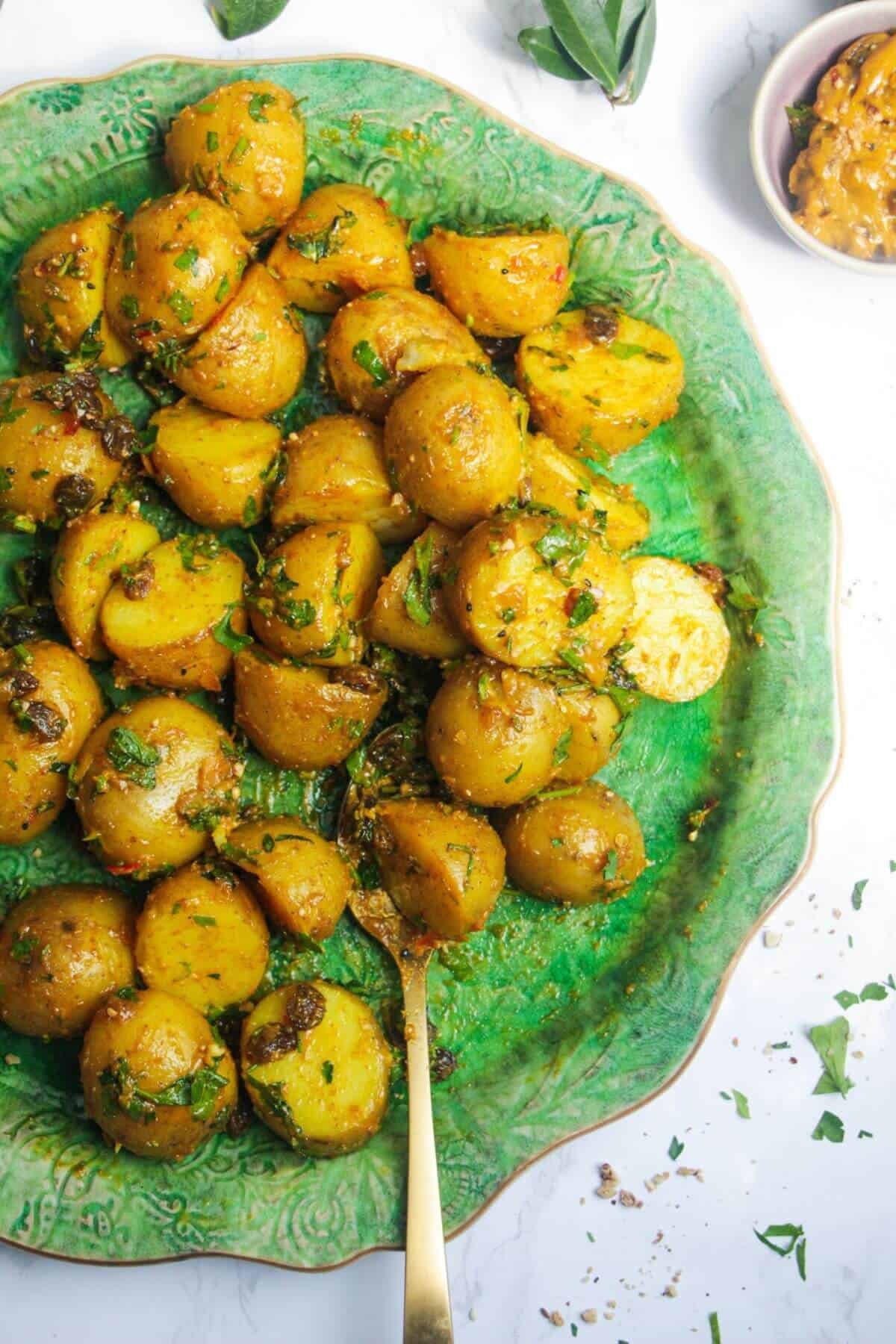 Coronation potato salad on a large green platter with a gold serving spoon and small bowl of coronation sauce on the side.