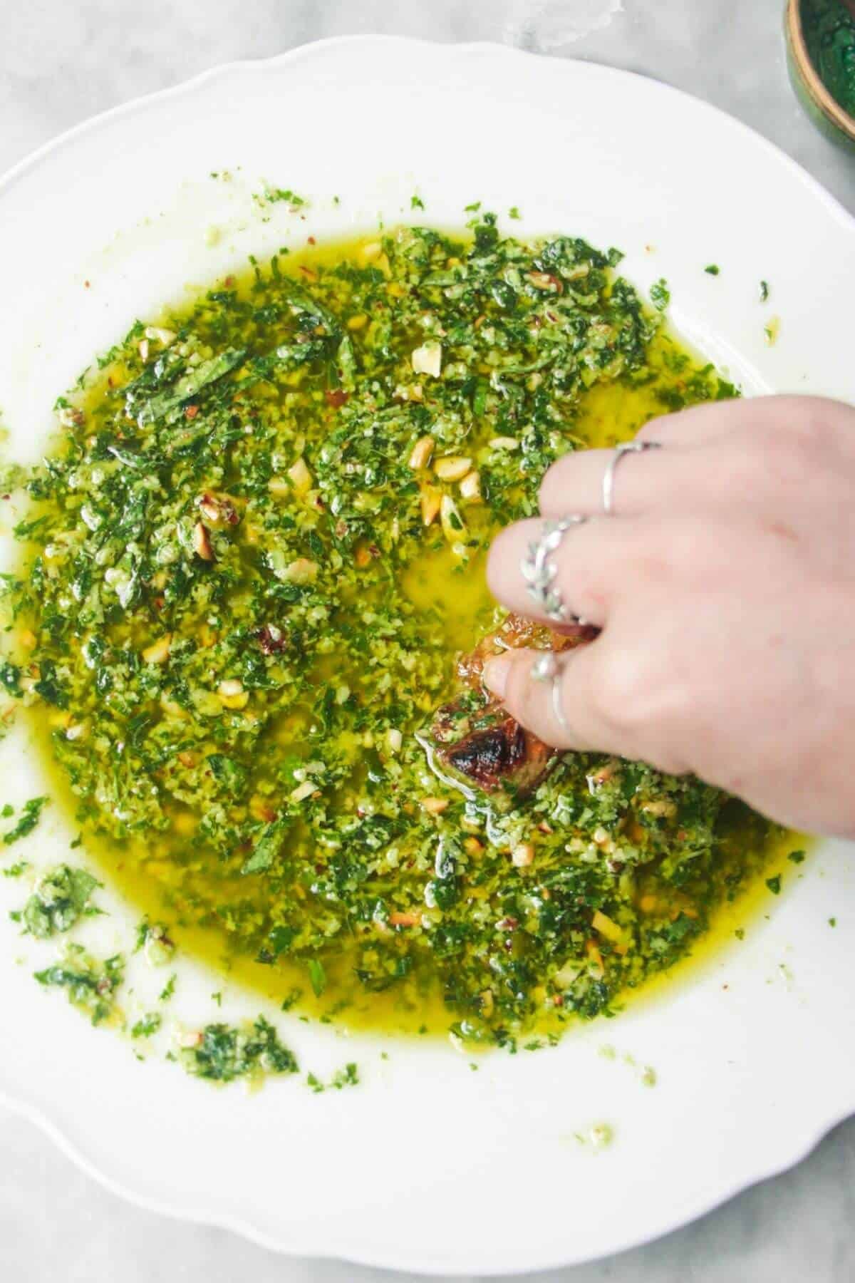 A hand sweeping bread through pesto dipping oil in a scalloped white plate.
