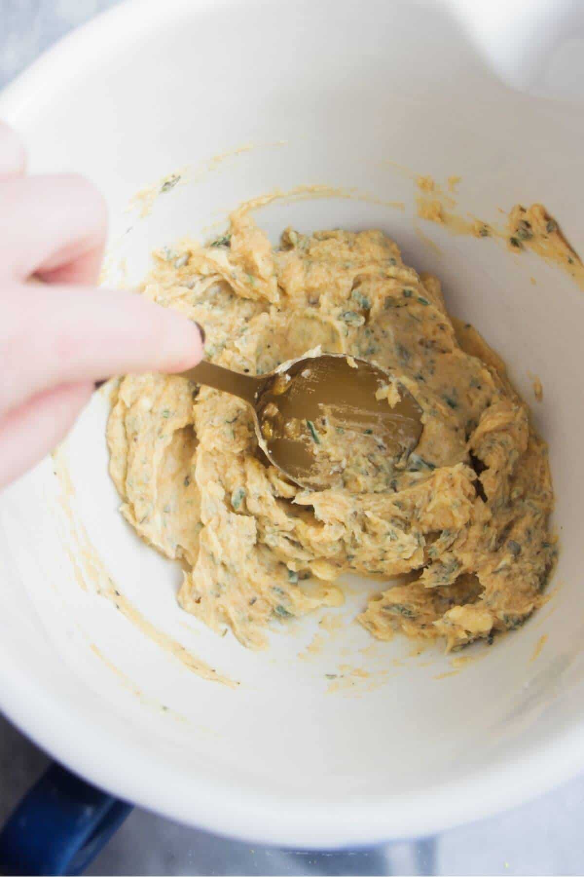 A gold spoon mixing butter, miso, honey and herbs in a white mixing bowl.