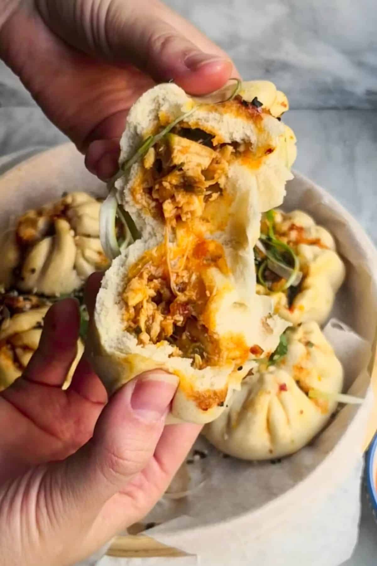 Two hands opening sticky chicken steamed bun with other cooked buns in the background.