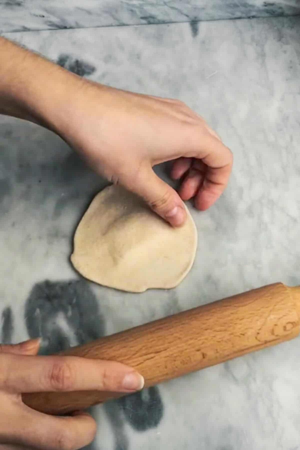 Hands about to pick up a rolled out circle of dough on a grey marble background.