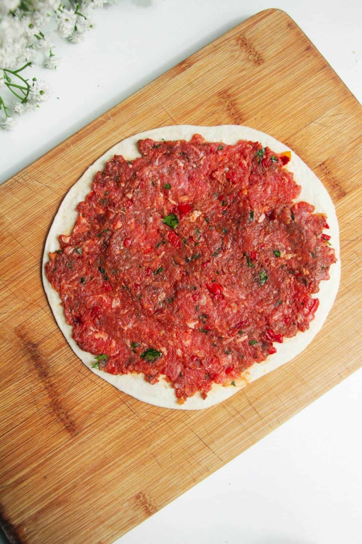 Lahmacun meat mixture spread onto a mini tortilla on a wooden board.