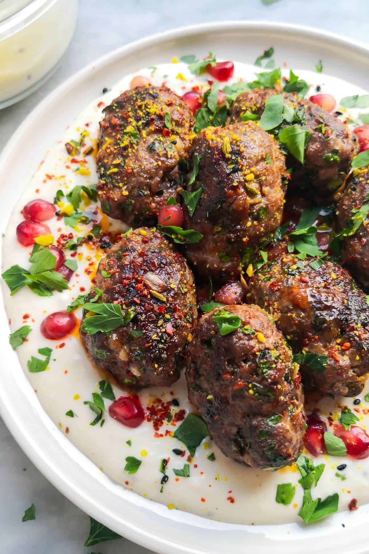 Lamb kofta sitting on yogurt tahini sauce, with pomegranate seeds, parsley, chilli flakes and dukkah on top with a bowl of yogurt tahini sauce in the background.