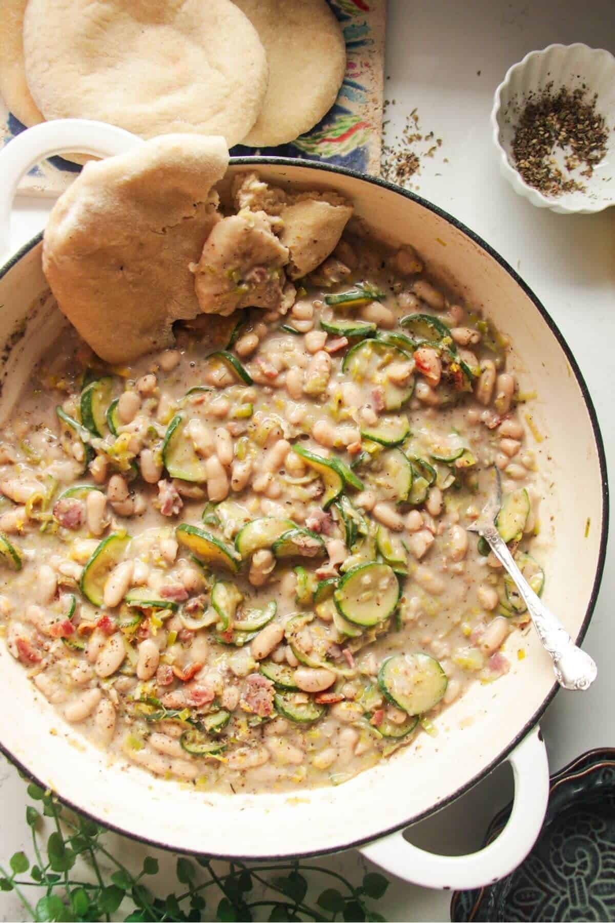 Creamy leek zucchini beans in a large pot with a silver spoon and pita bread on the side.