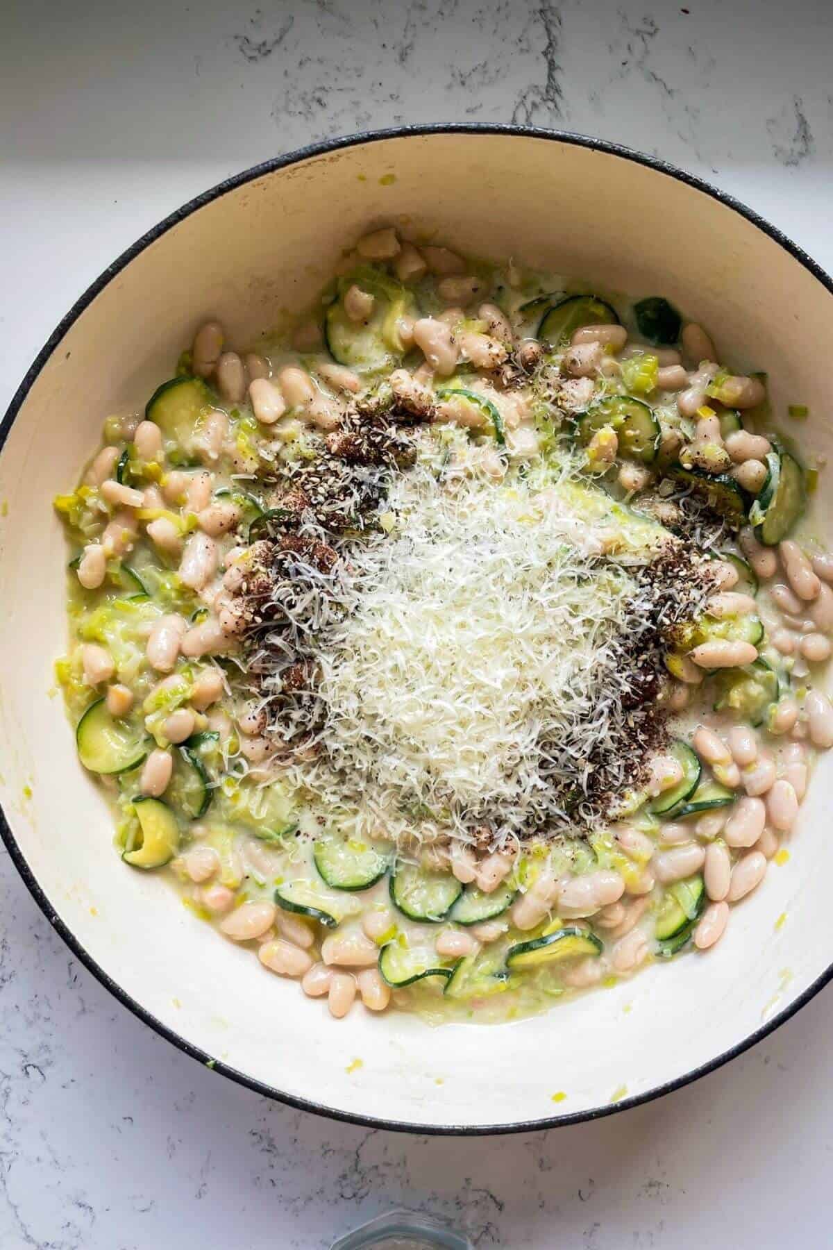 Za'atar and grated parmesan on top of white beans, zucchini and leeks in a large pot.