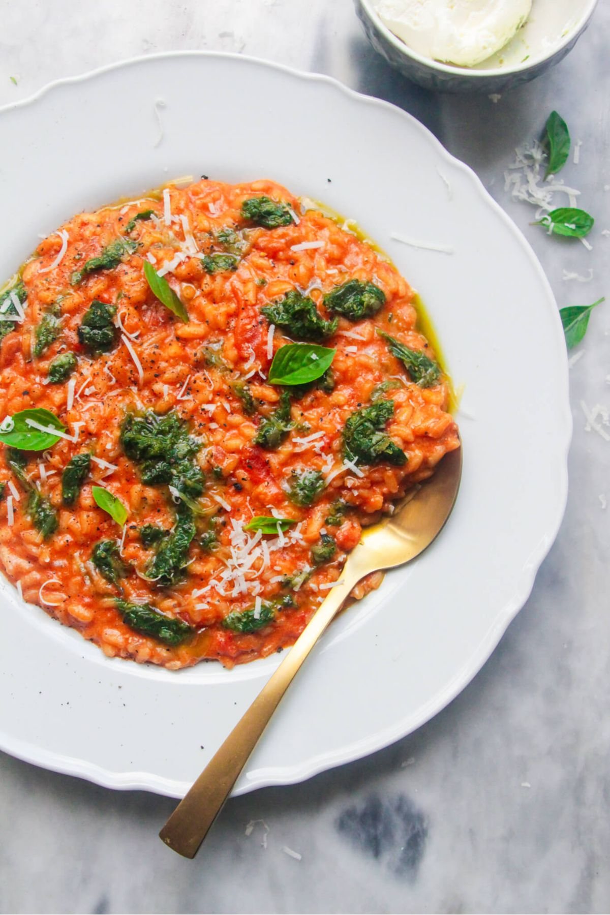 Tomato mascarpone risotto in a large white plate with dollop of green pesto on top, with a gold spoon in the risotto and bowl of mascarpone in the background.