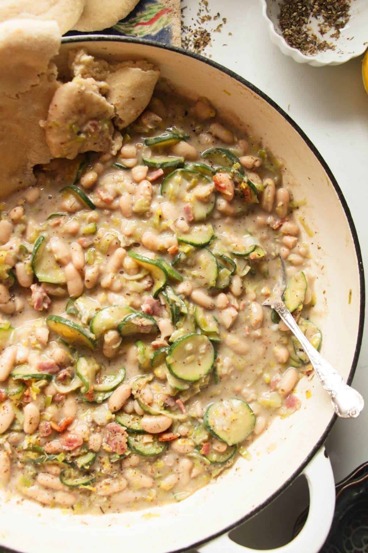 Creamy leek zucchini beans in a large pot with a silver spoon and pita bread on the side.