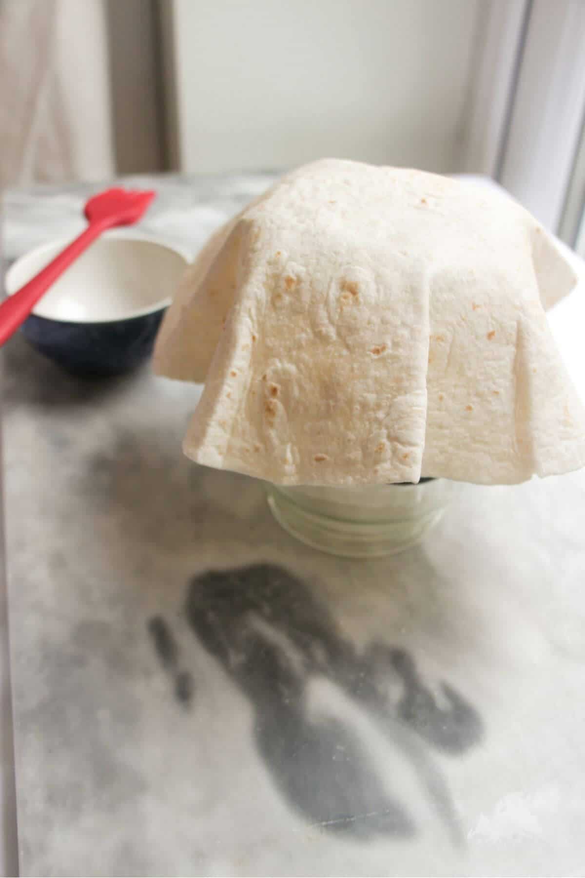 Tortilla draped over a large glass jar with small bowl of oil in the background.