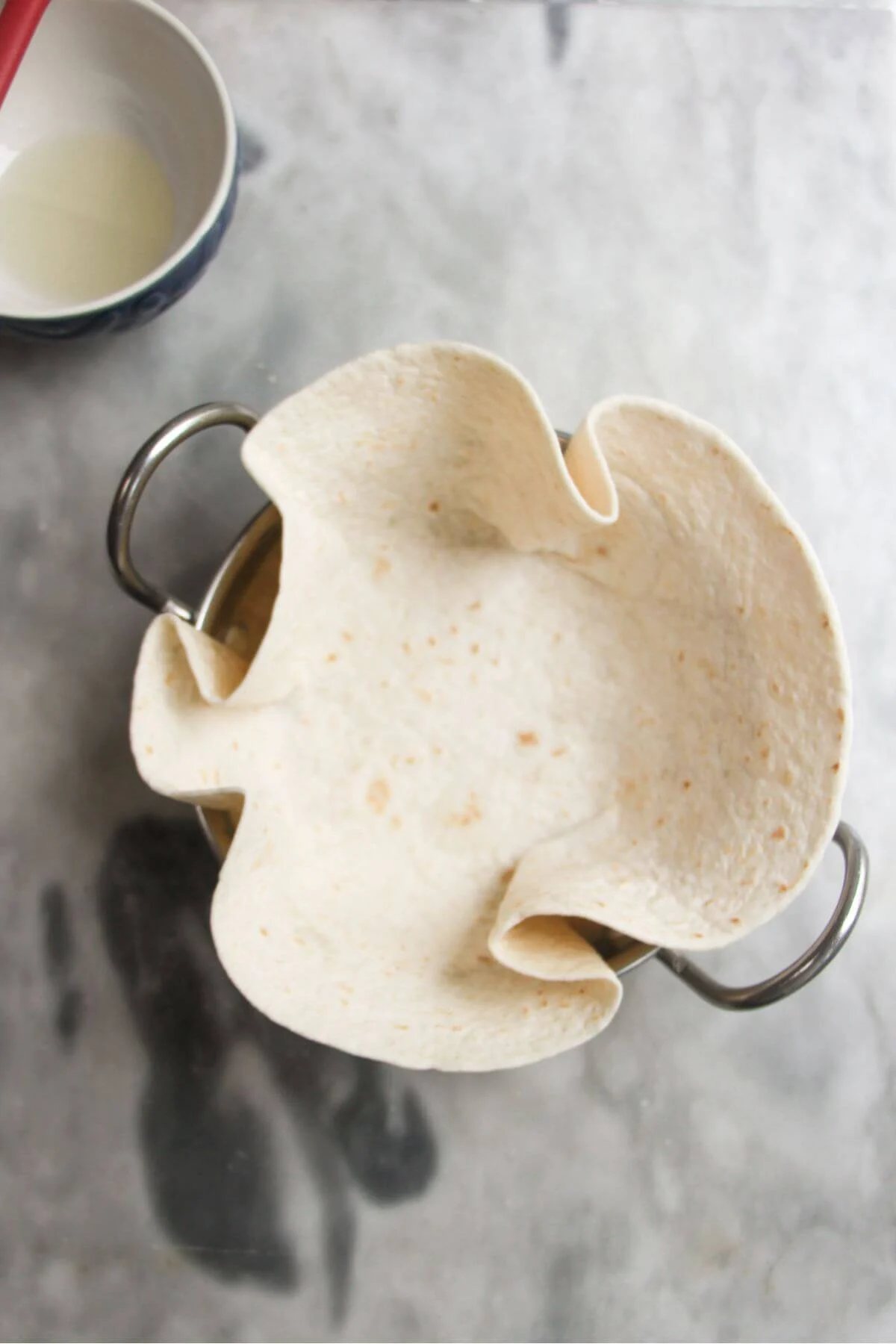 Tortilla pressed into a small silver bowl with small bowl of oil in the background.
