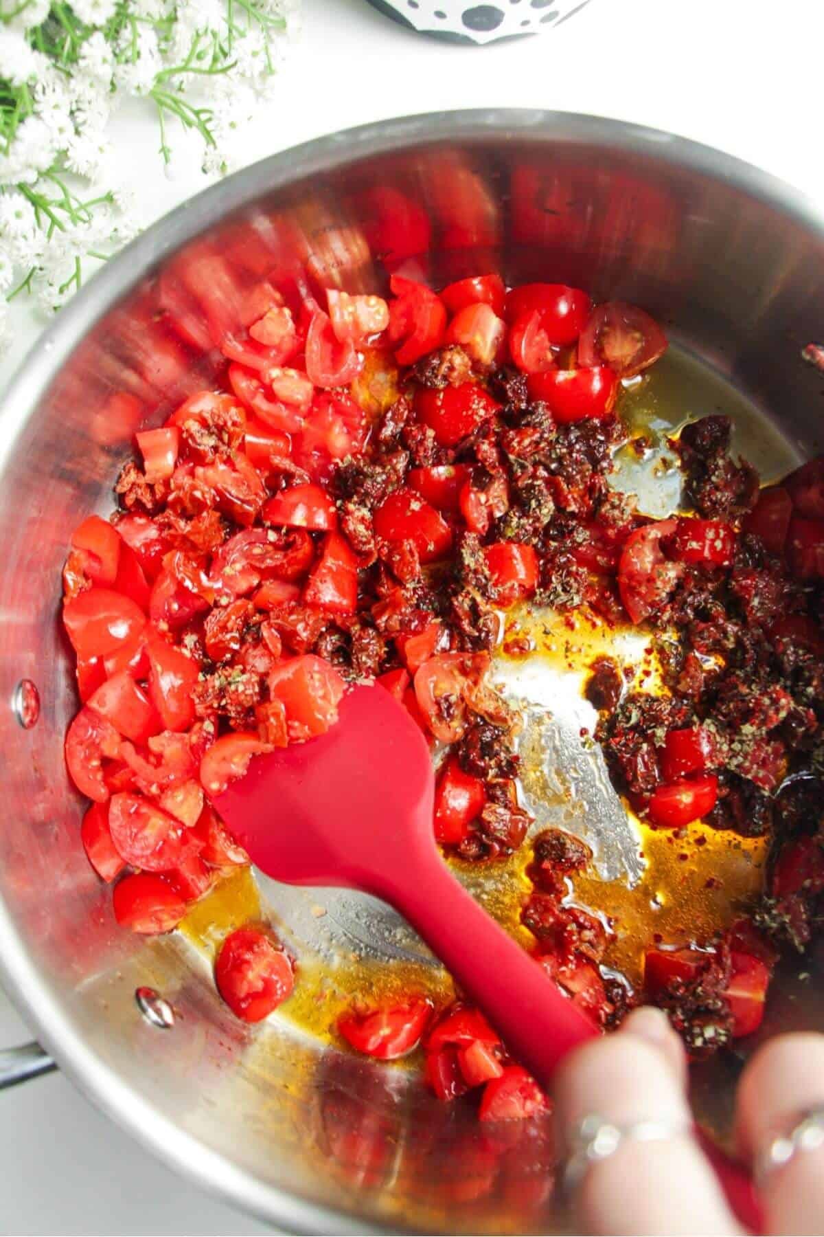 Red spatula stirring tomatotes in a large stainless steel pan.