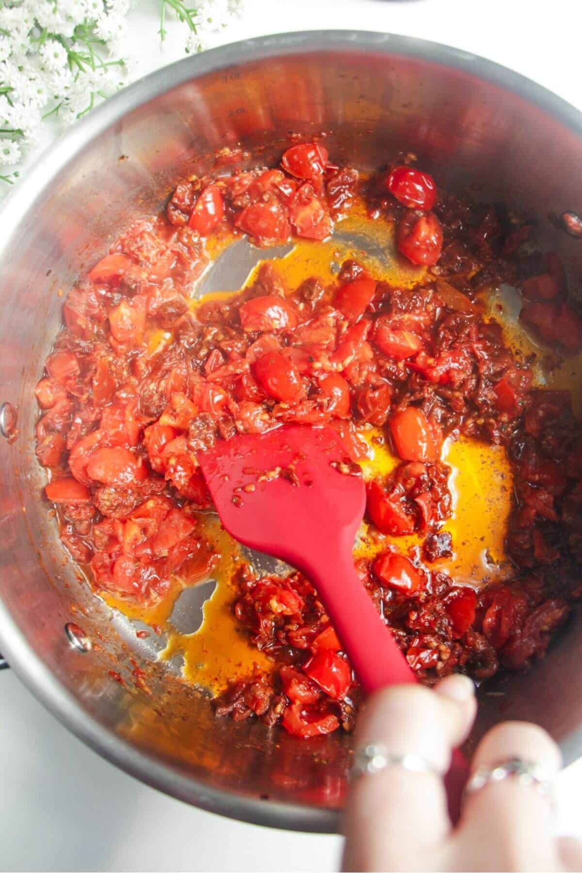 Red spatula stirring tomatotes in a large stainless steel pan.