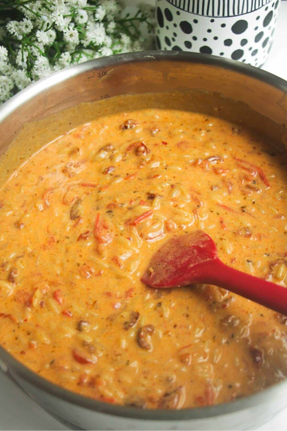 Red spatula stirring creamy tomato orzo in a stainless steel pot.
