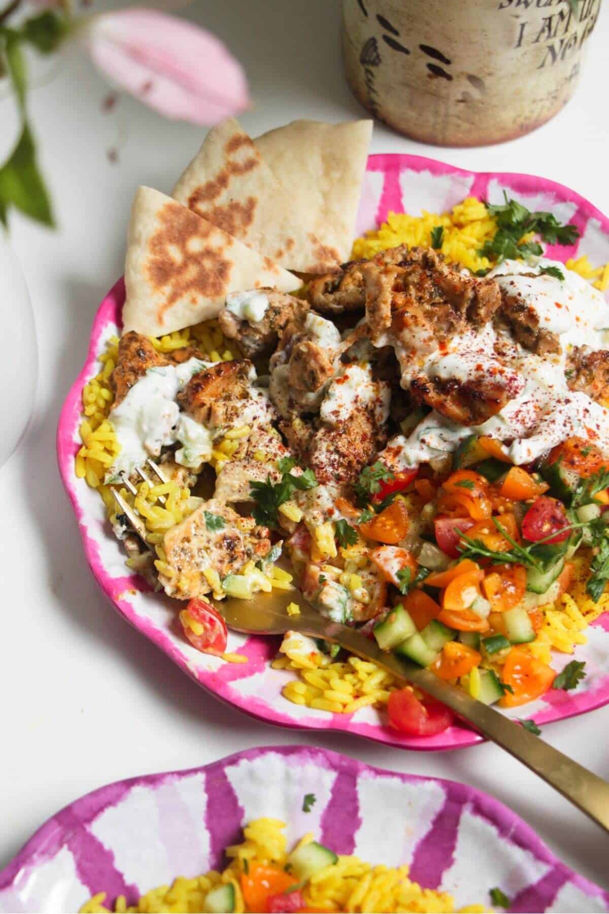 Chicken, salad and rice on a pink and white striped plate with pita bread on the side.