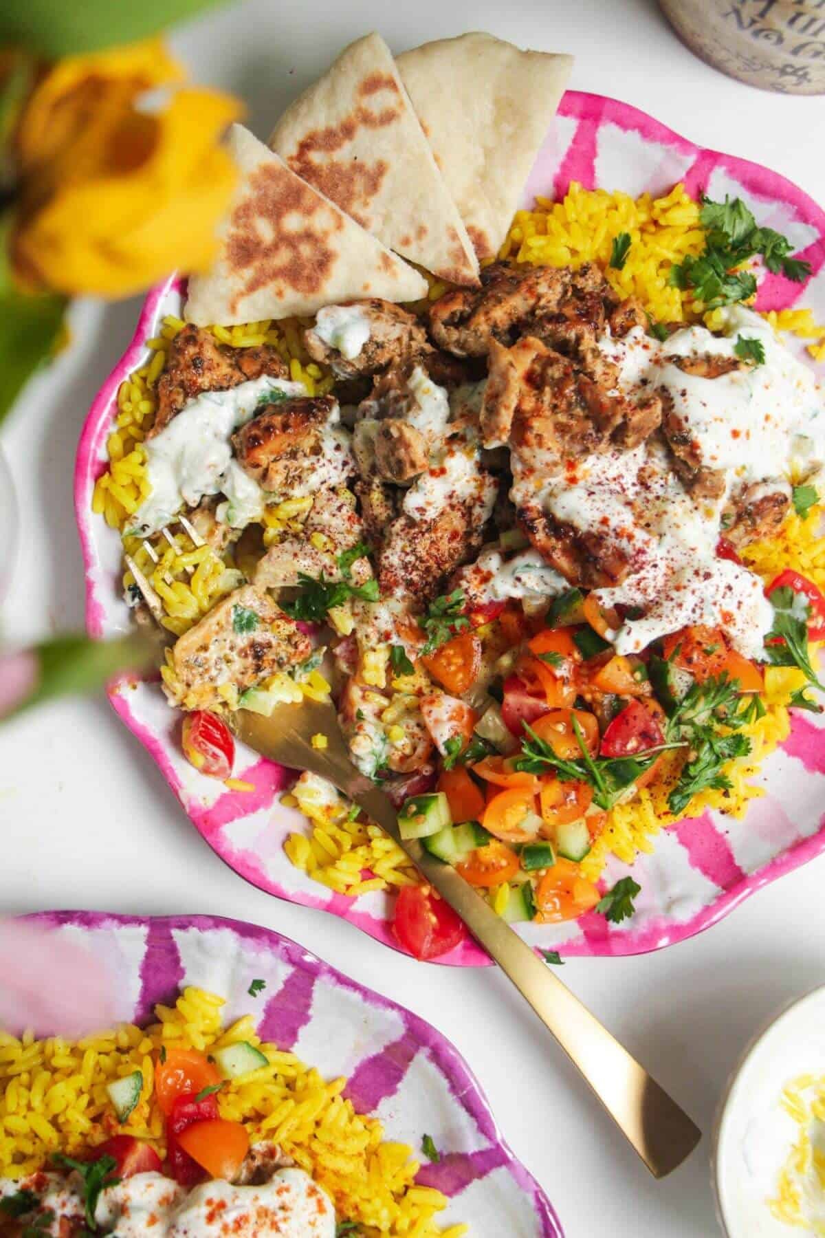 Chicken, salad and rice on a pink and white striped plate with pita bread on the side.