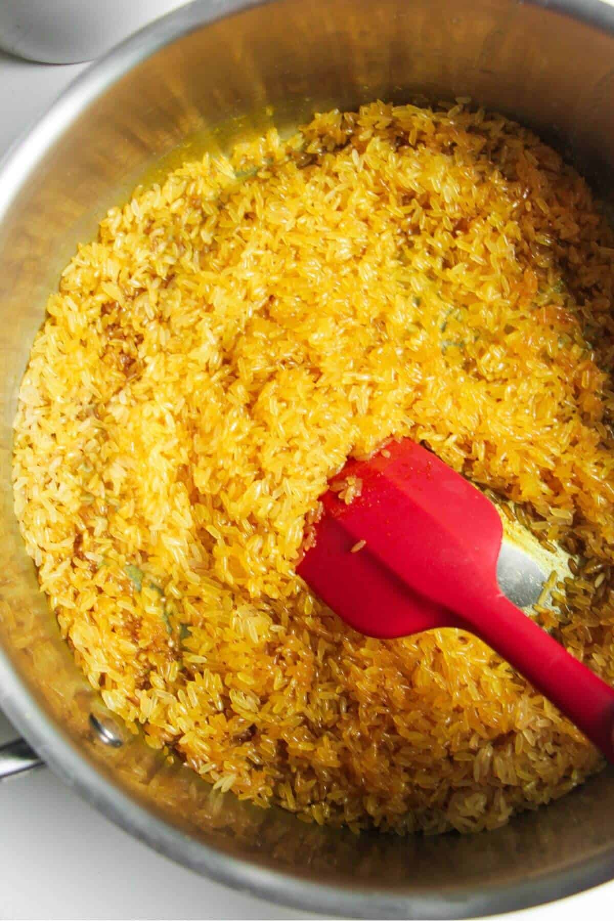 Red spatula stirring rice through spices in a large pot.