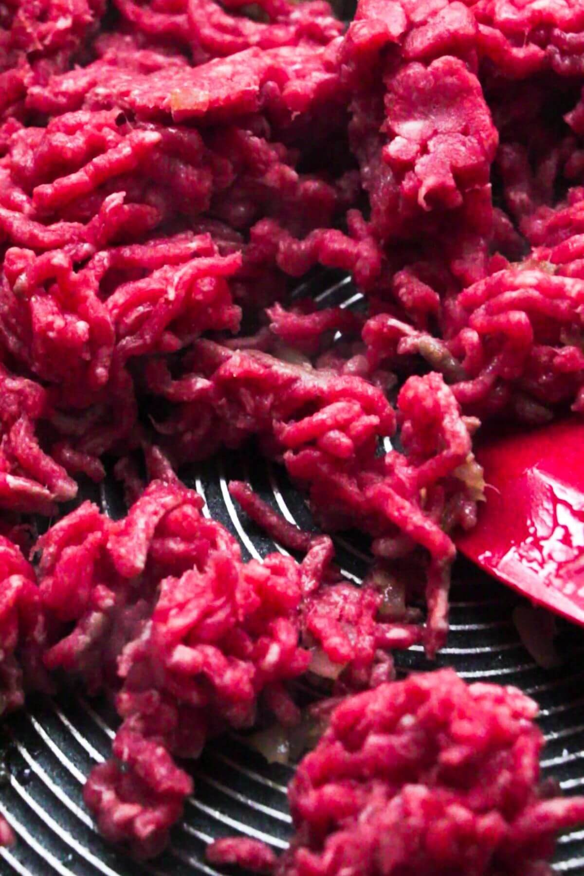 Close up of raw mince being added to a large pan and being stirred with a red spatula.