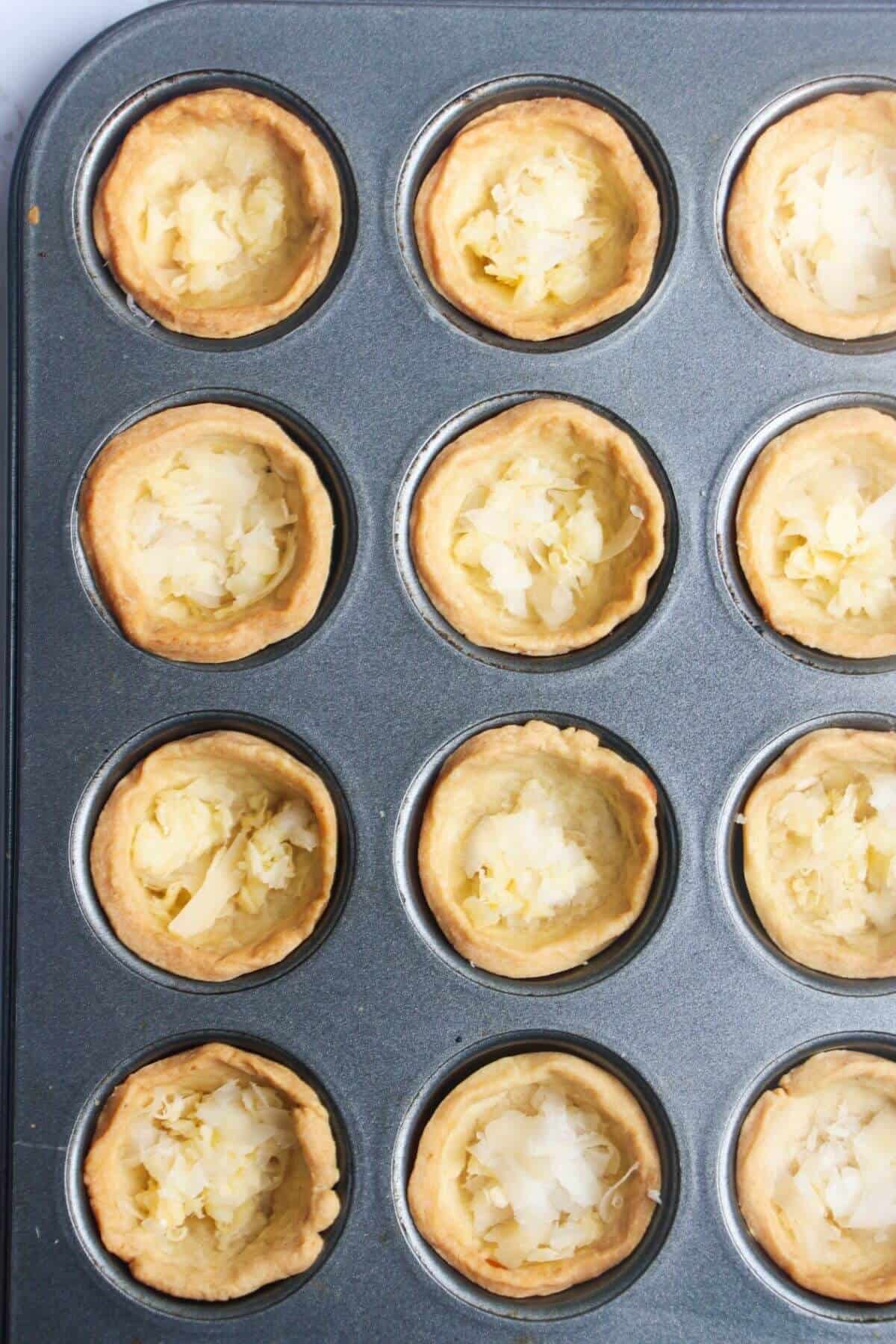 Grated cheese on the base of mini pastry cases in a mini muffin tin.