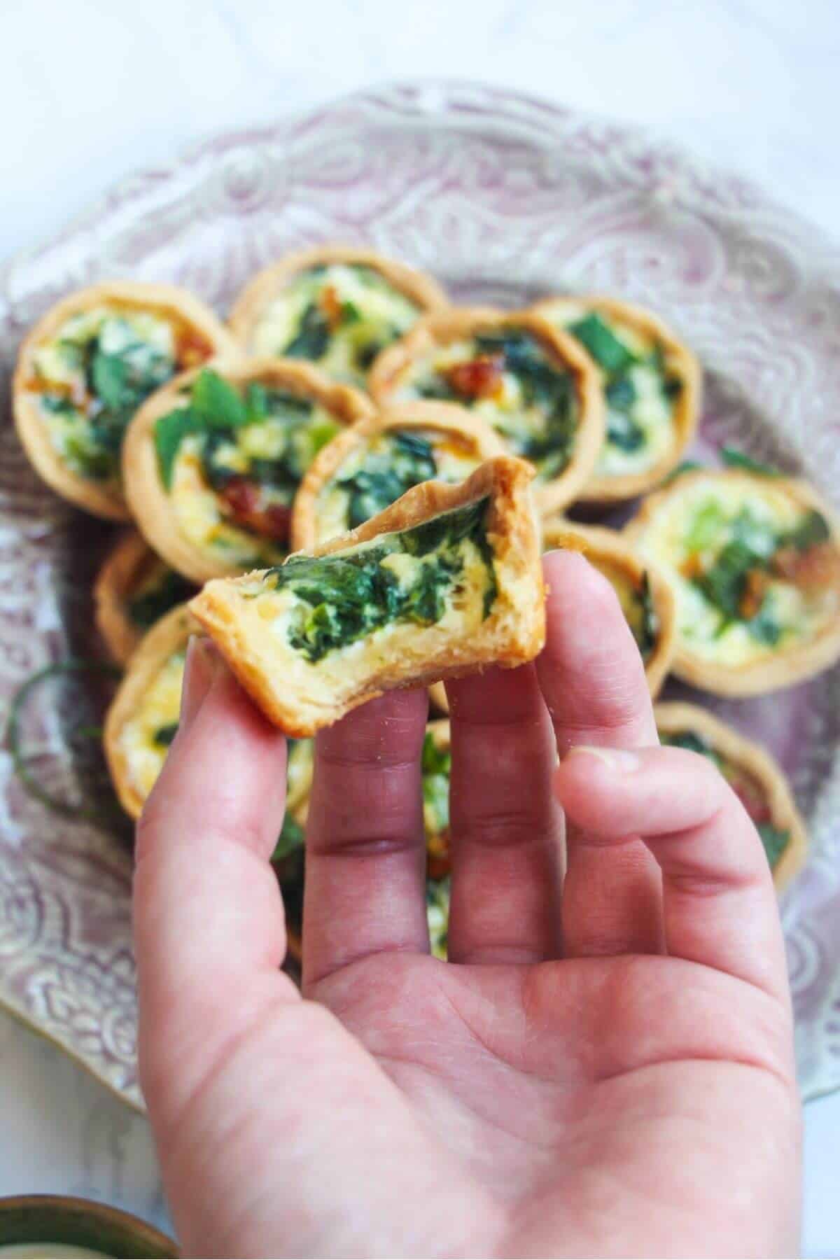 Hand holding a bitten mini quiche with another plate of quiches in the background.