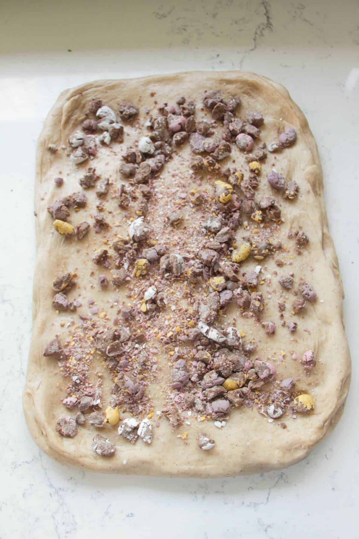 Crushed mini eggs scattered on top of hot cross bun dough stretched out into a large rectangle.