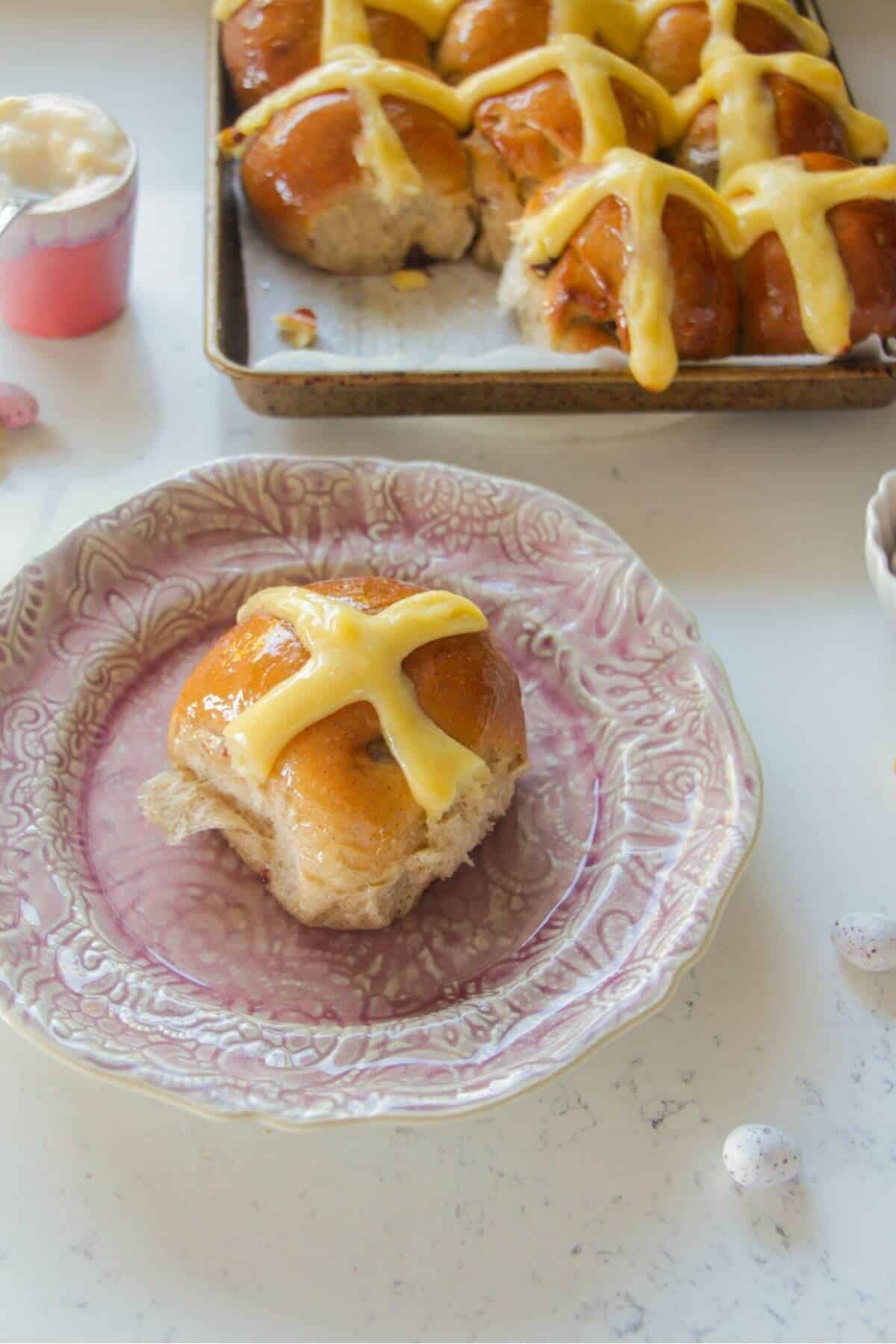 Close up of a mini egg hot cross bun on a small pink plate with more hot cross buns in the background.