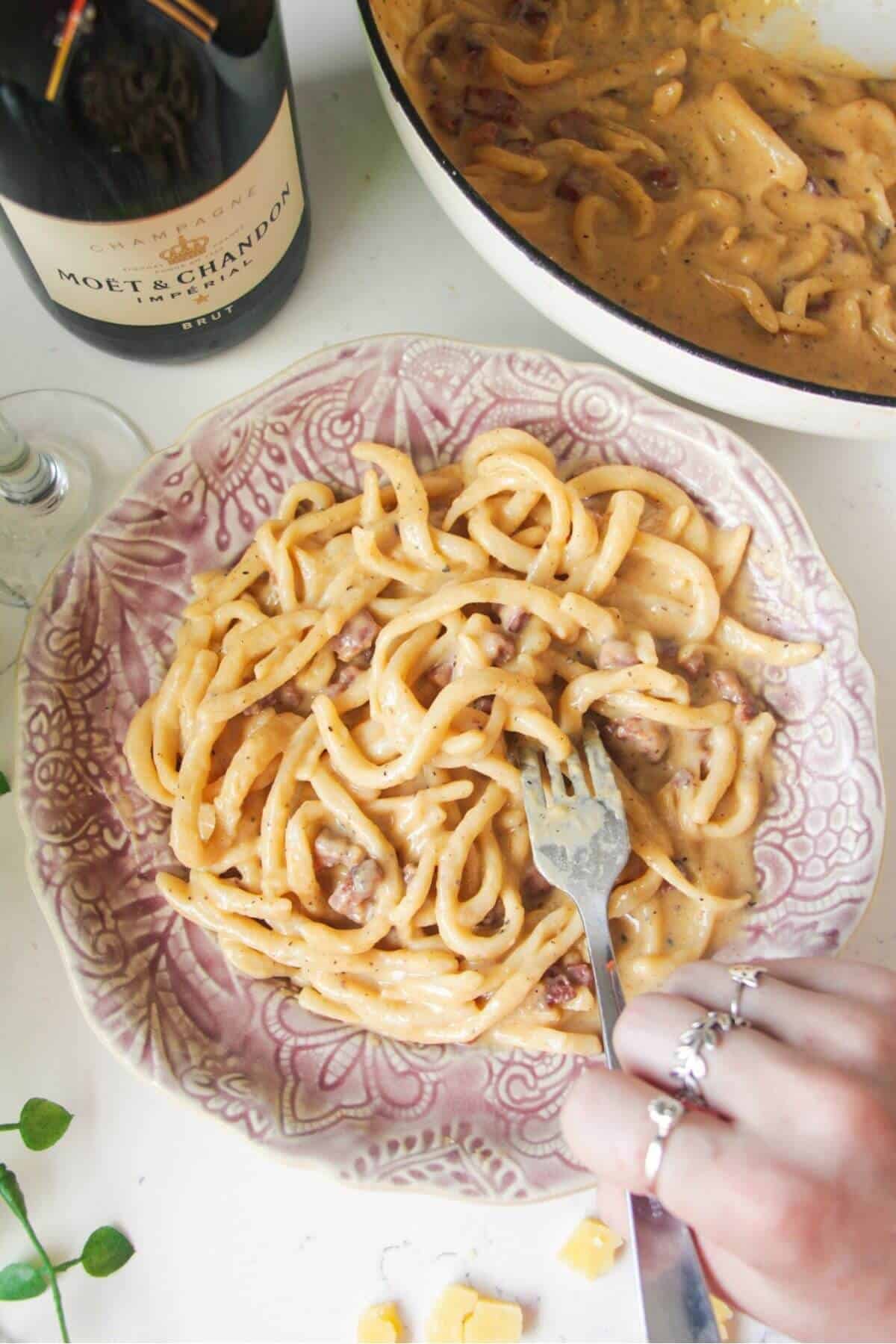 Hand holding fork swirling through spaghetti carbonara on a small pink plate.