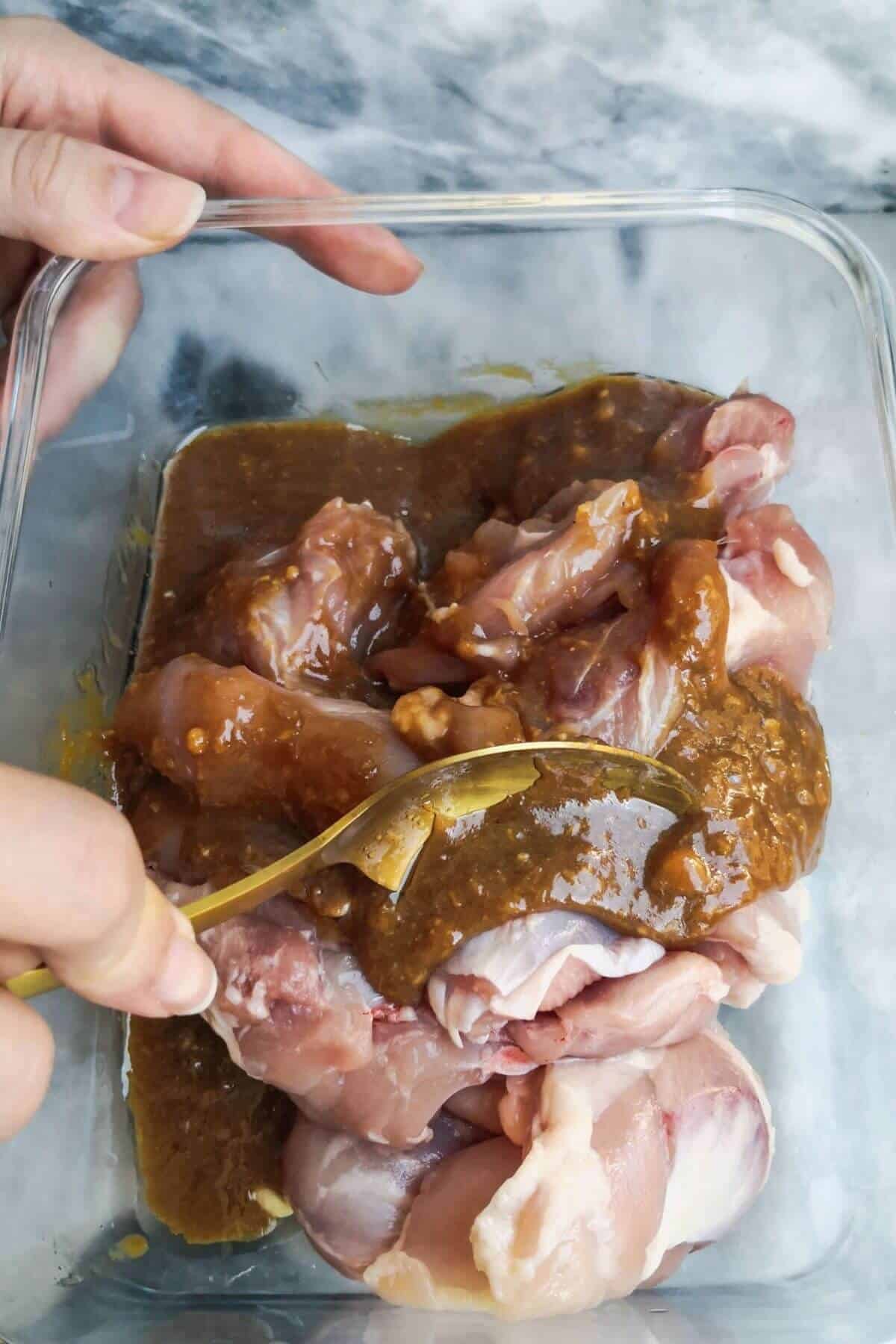 Gold spoon mixing chicken thighs with the miso marinade in a glass container on a grey marble background.