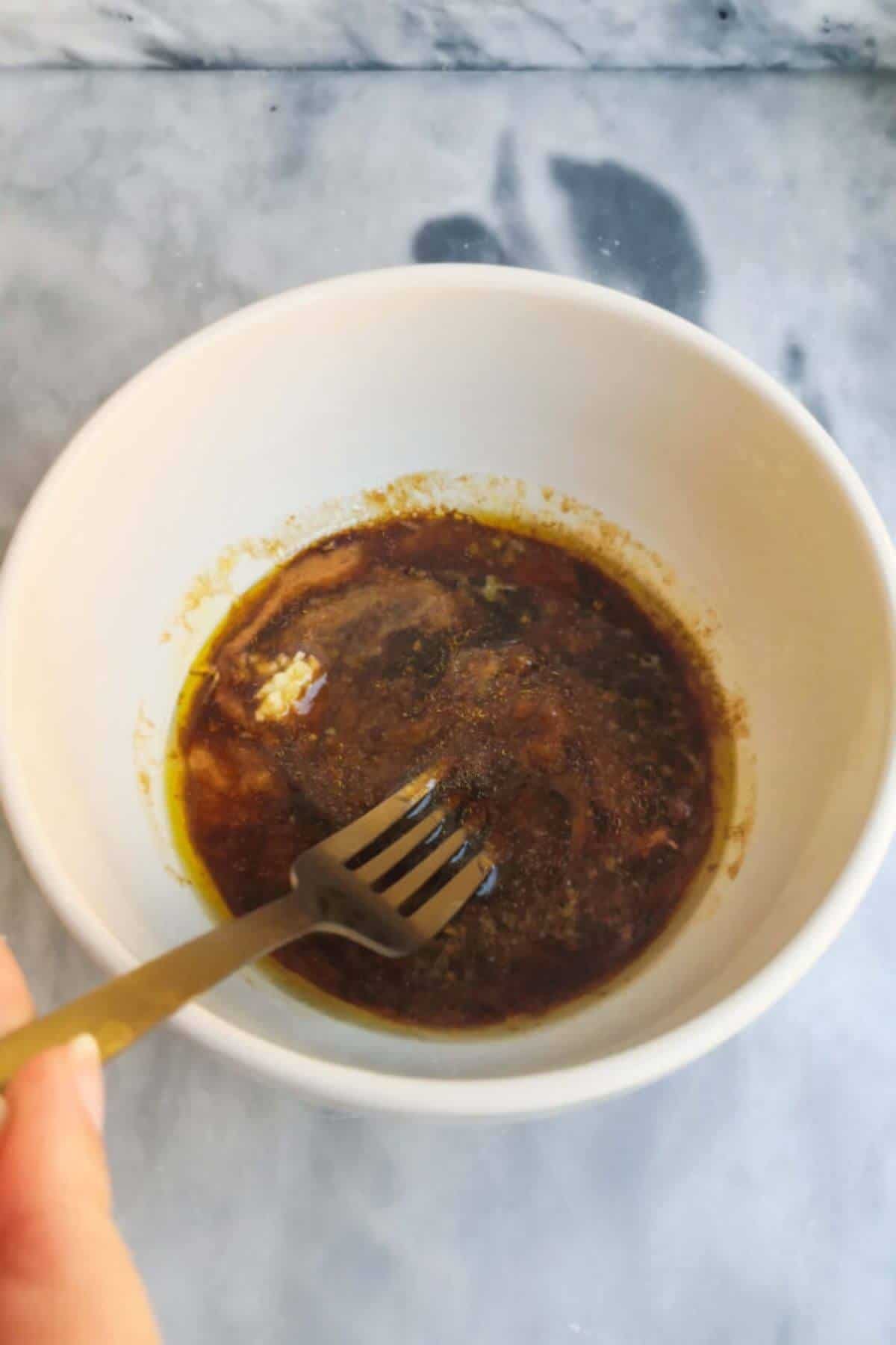 A gold fork mixing miso marinade in a small white bowl on a grey marble background.