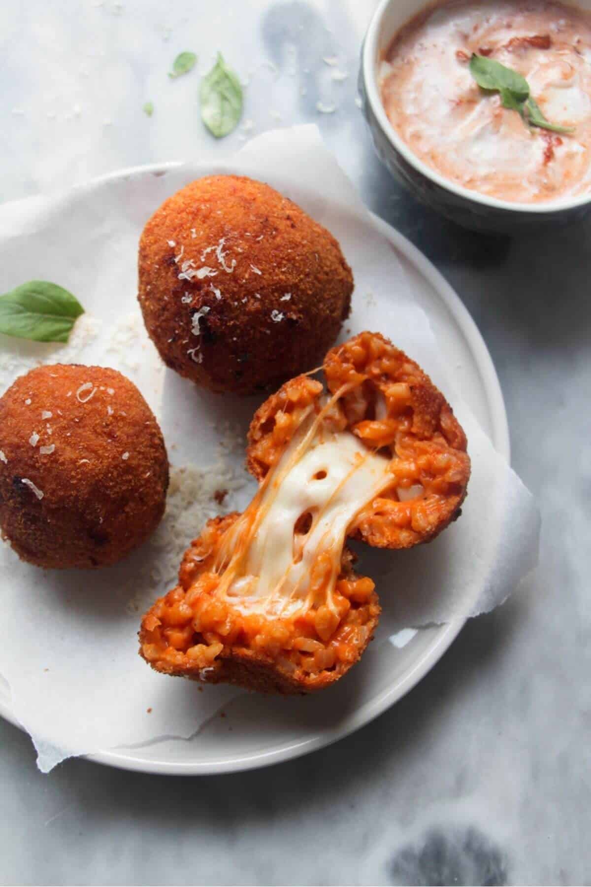 3 whole arancini on a small plate with a open arancini with cheese spilling out, with aioli with pesto on the side in a small bowl.