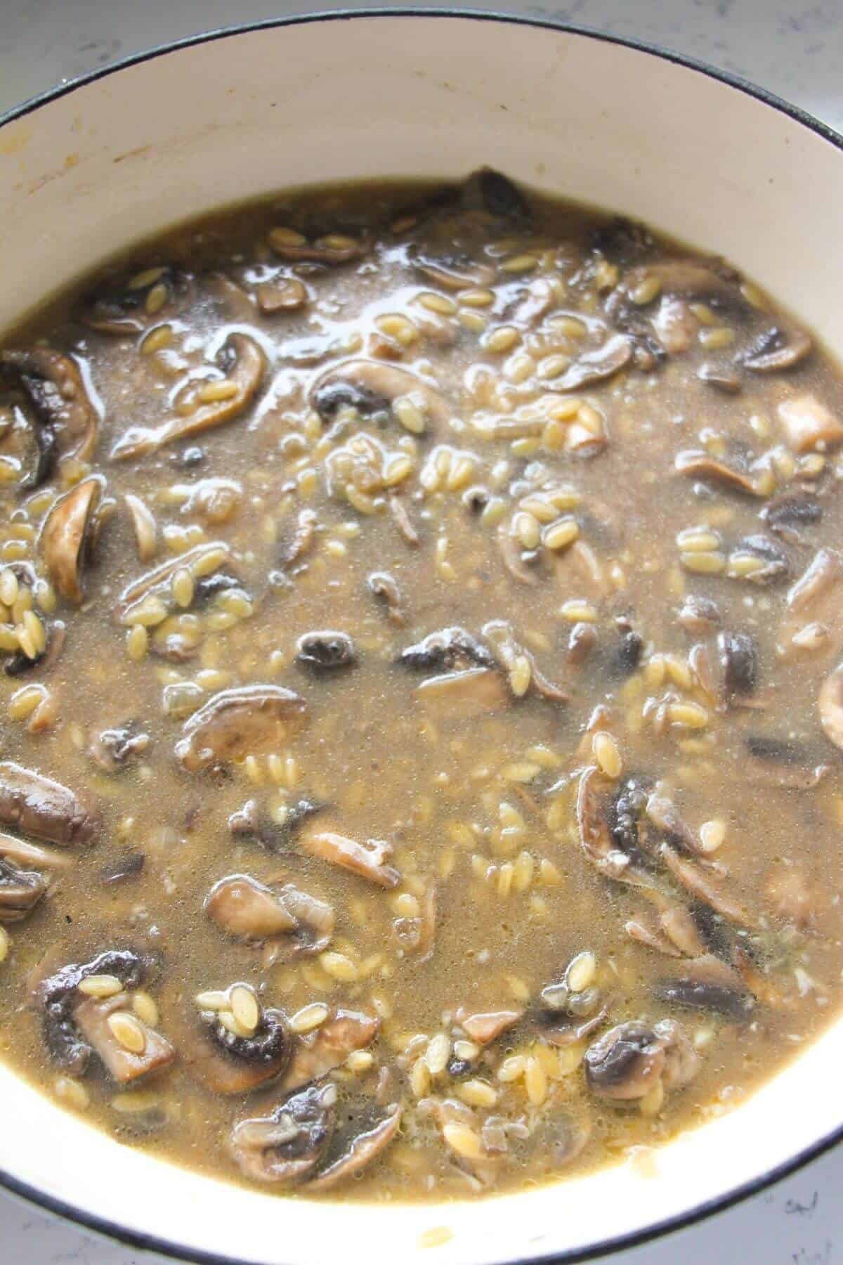Stock added to mushrooms and orzo in a large white skillet.
