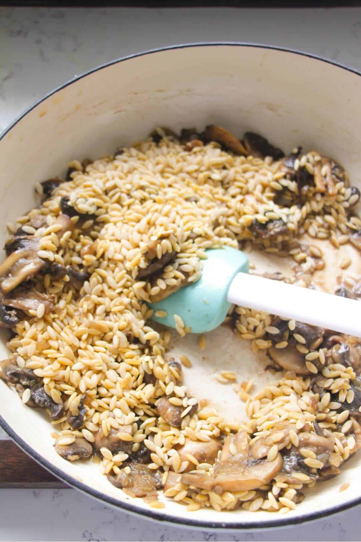Orzo being stirred through mushrooms in a large white skillet.