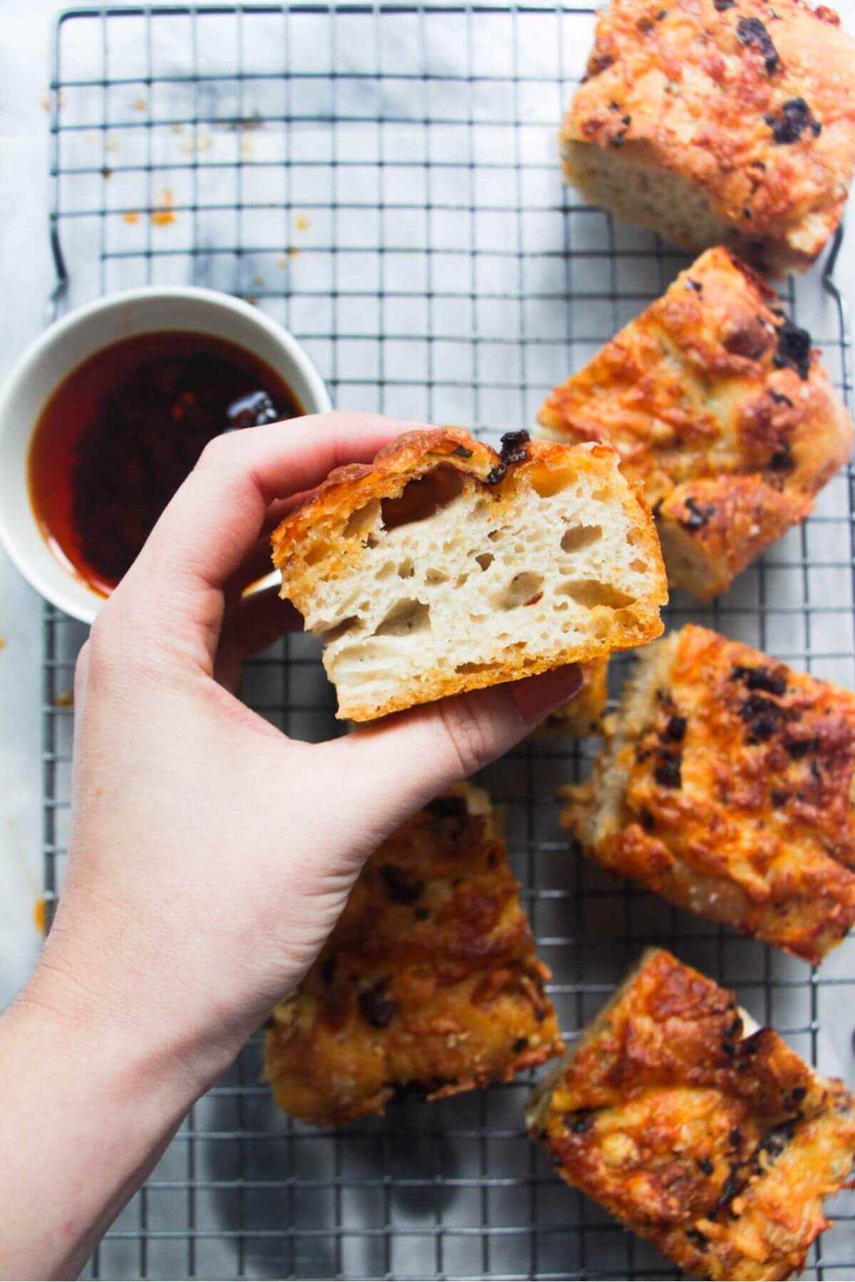 Hand holding up a piece of focaccia with chopped focaccia in the background.