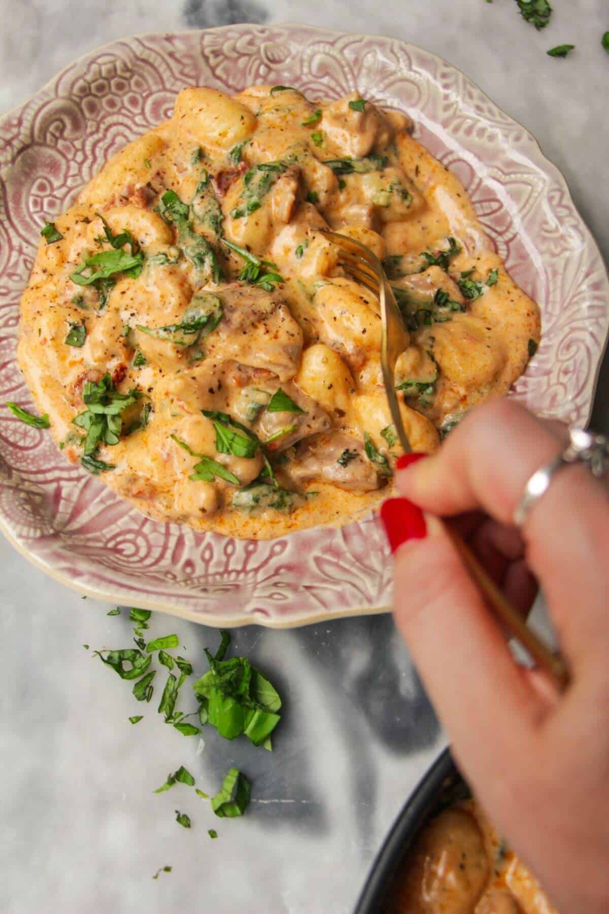 Hand holding a fork, with creamy chicken gnocchi in a pink plate.