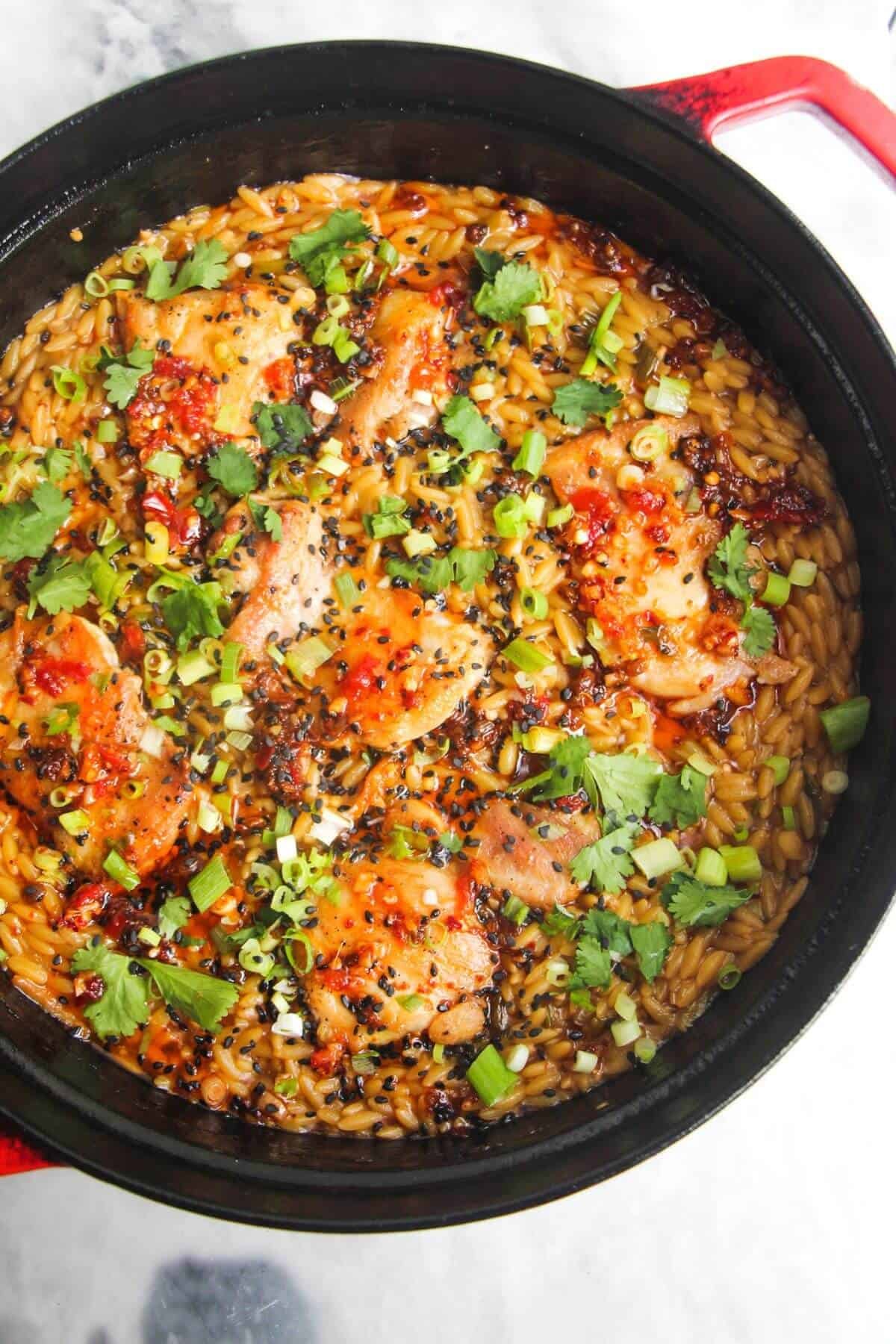 Cilantro, scallions and chilli oil drizzled on top of chicken thighs in orzo in a large black pan.