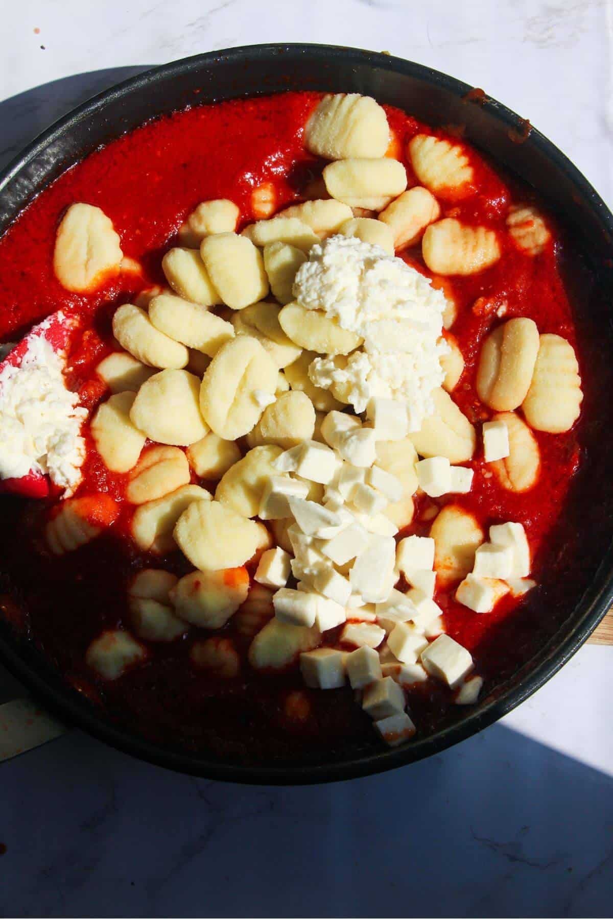 Gnocchi and cubed mozzarella being added to a frying pan with tomato sauce in it.