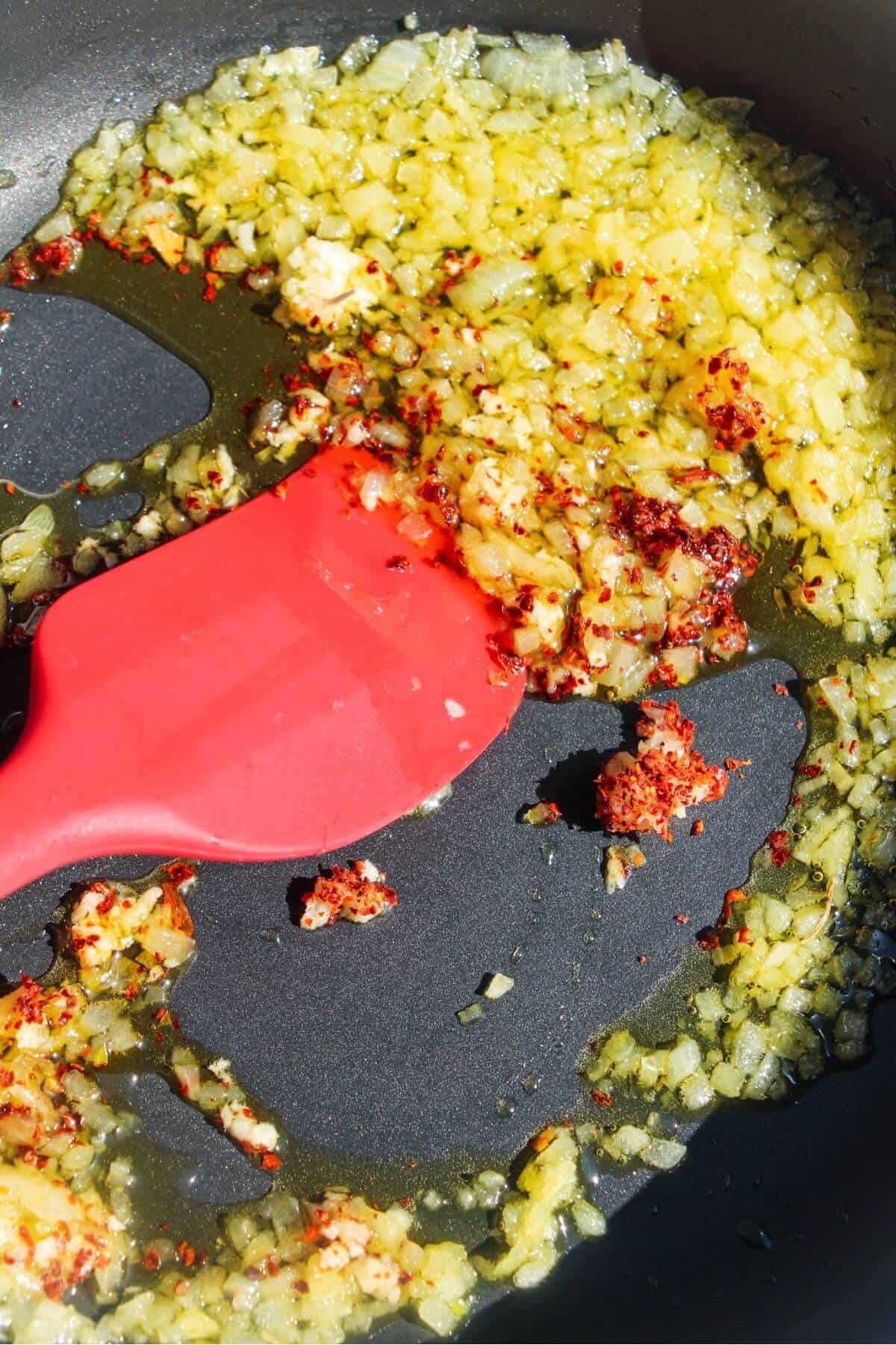 Red spatula stirring diced onion, garlic and red pepper flakes in a small frying pan.