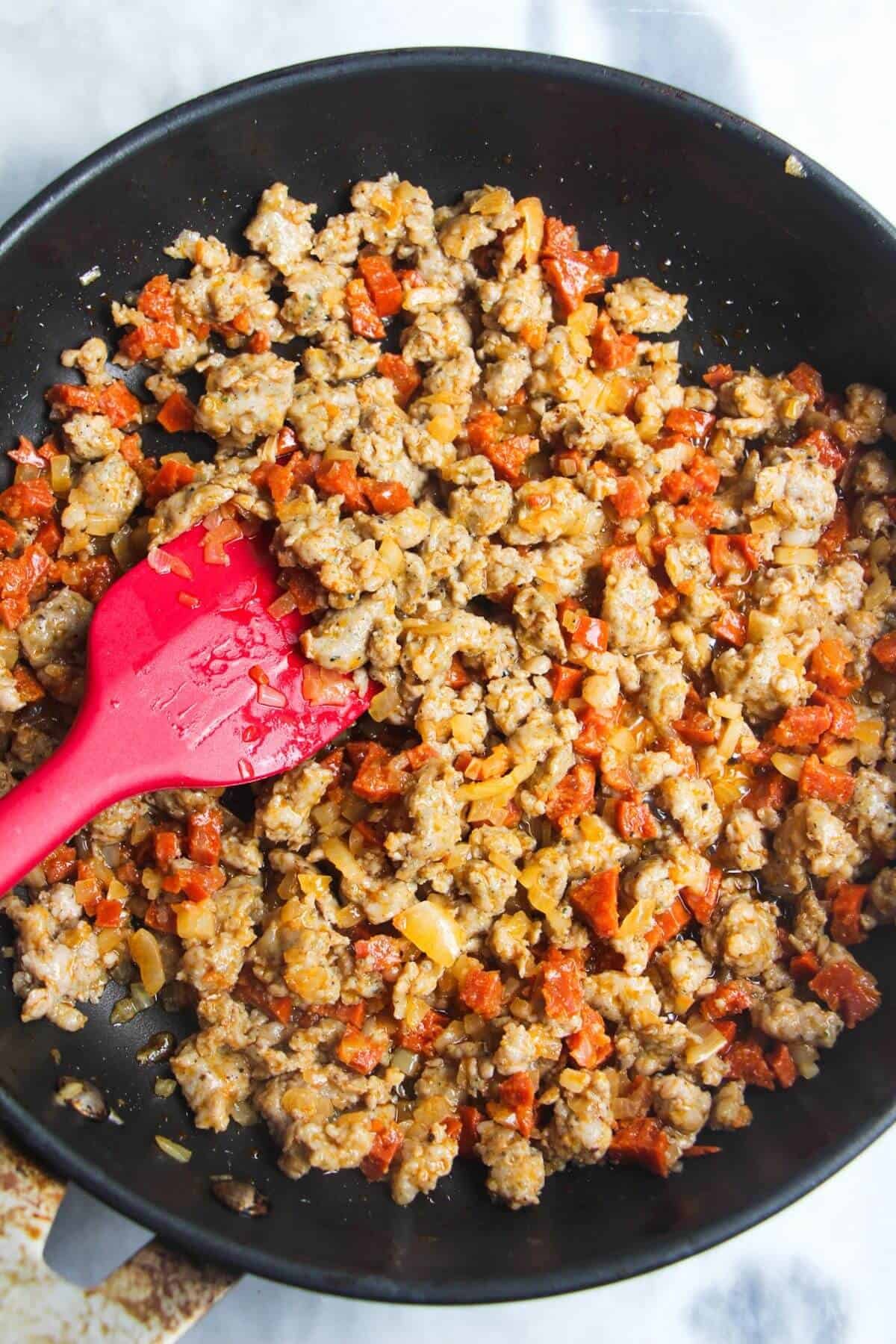 Red spatula stirring cooked sausage meat in a pan.