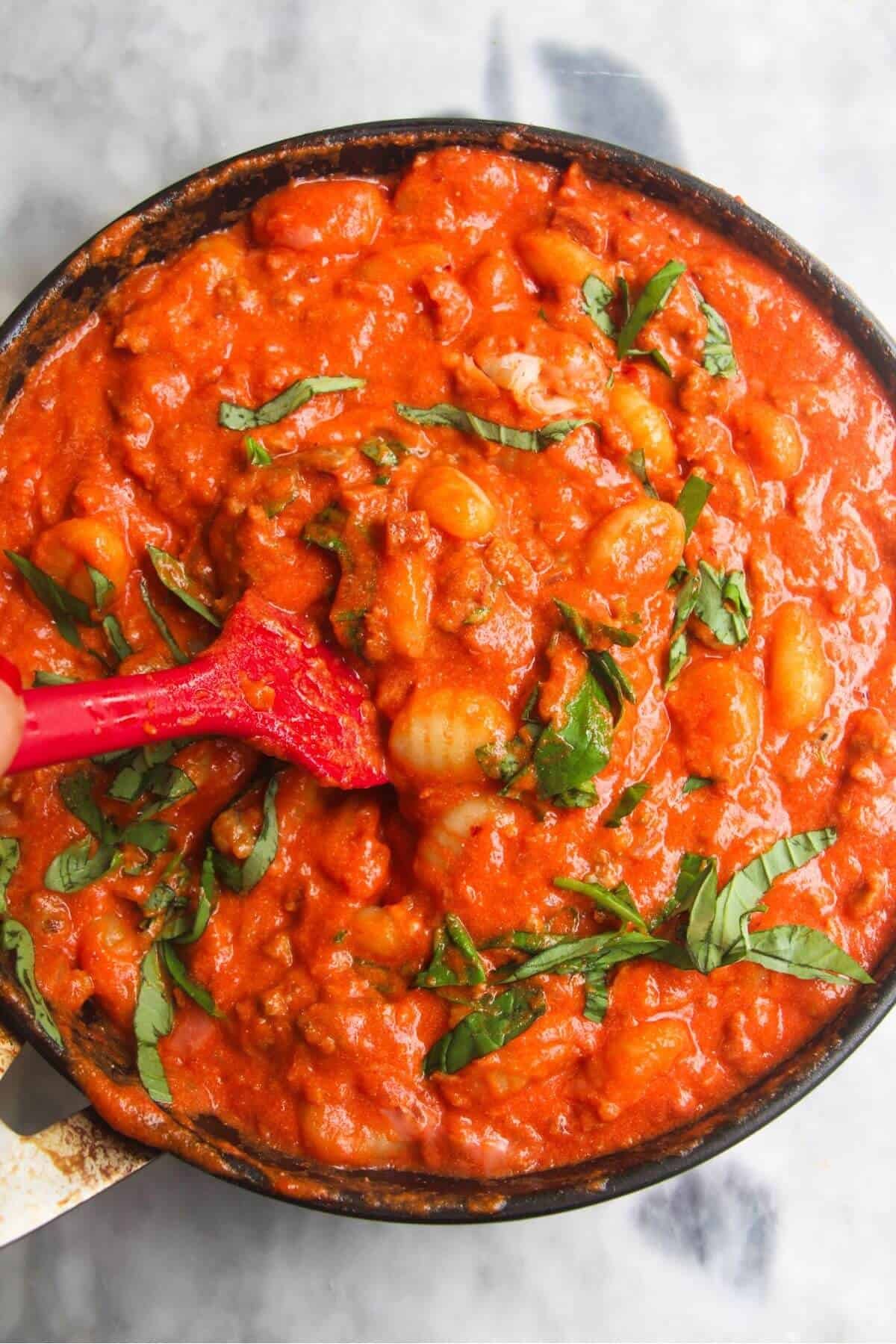 Red spatula stirring basil through creamy tomato gnocchi.