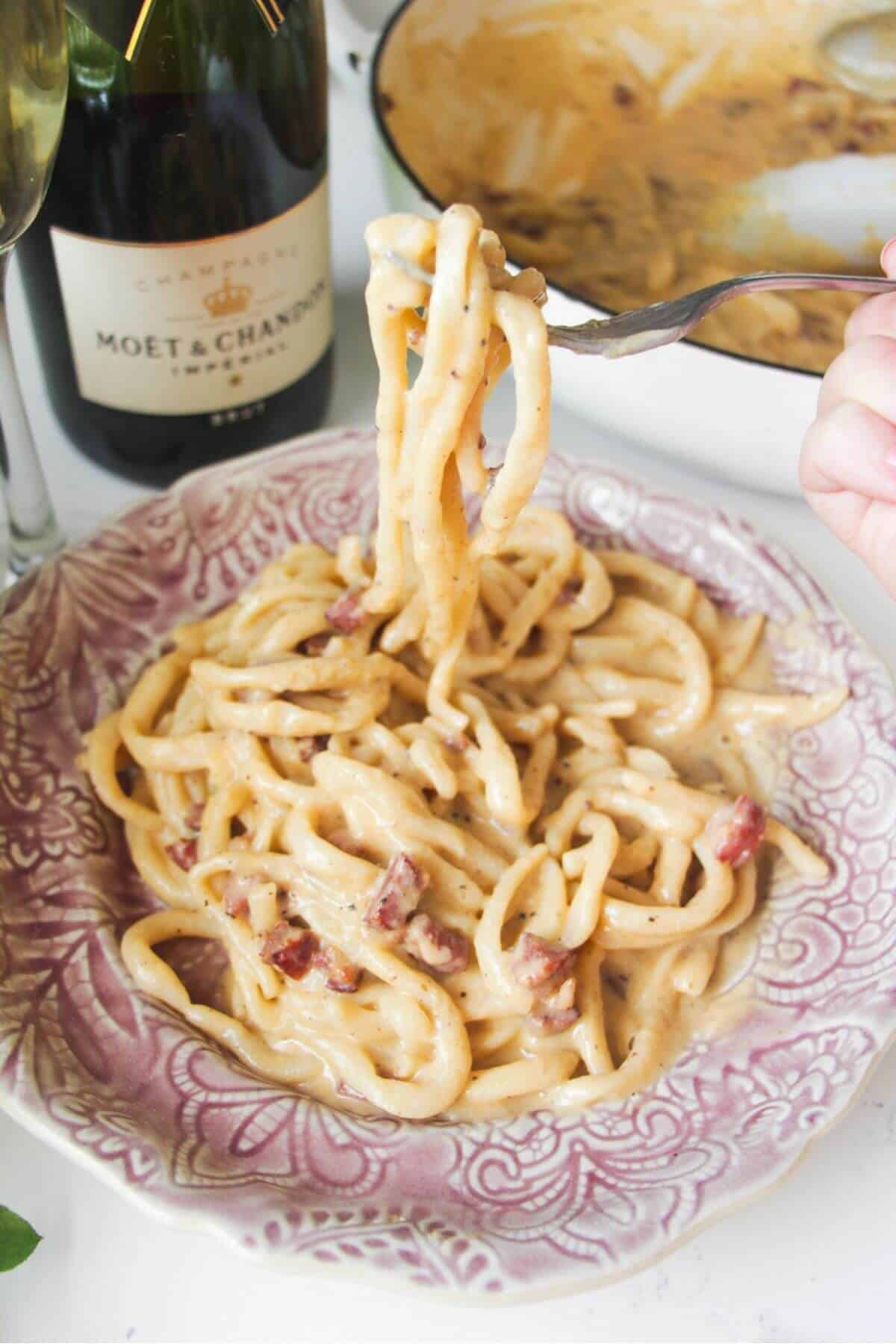 Fork holding up strands of aspaghetti, with a plate of carbonara in the foreground.