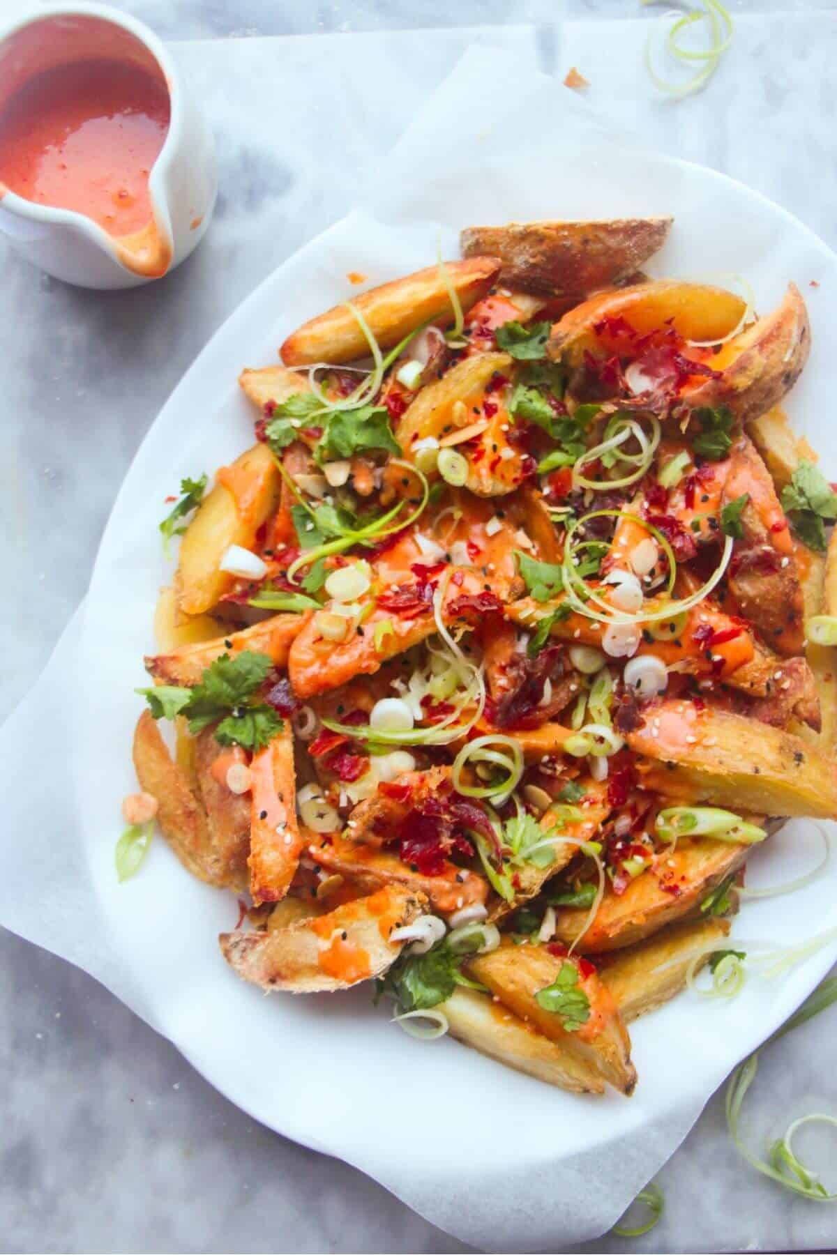 Loaded potato wedges on a large oval white plate with small jug of gochujang bechamel on the side.