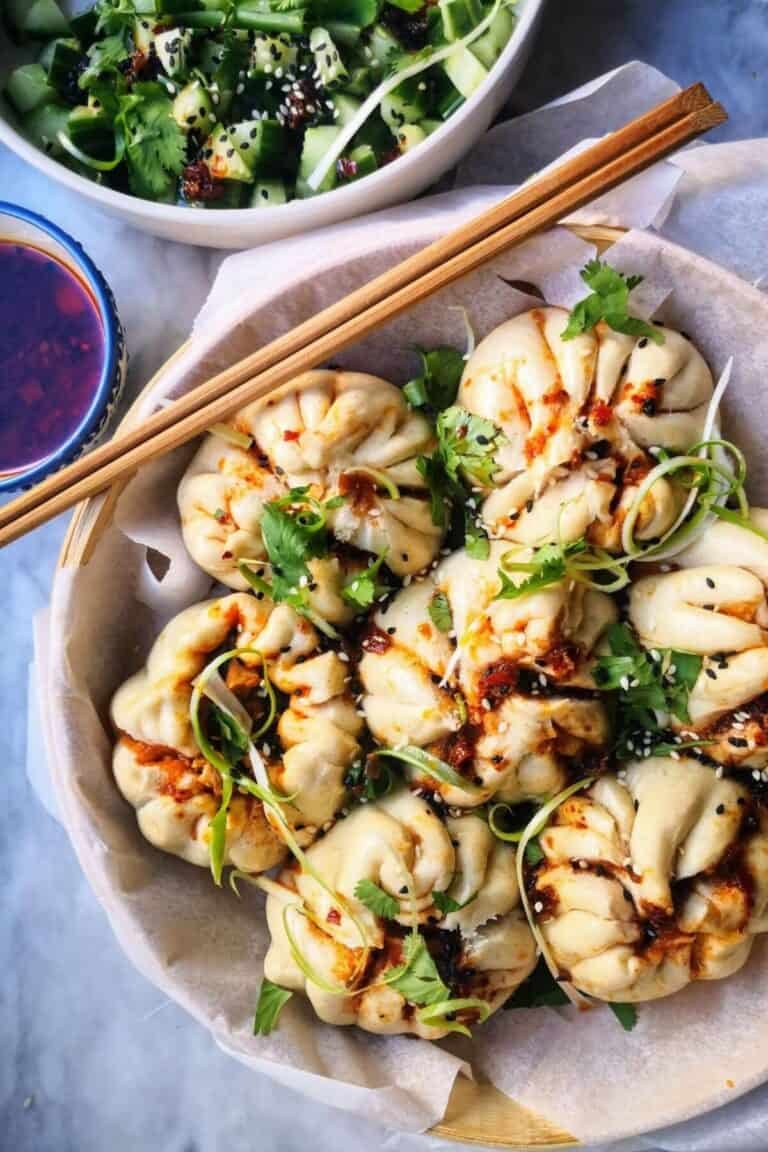 Sticky Korean chicken steamed buns in a bamboo steamer, with cucumber salad and chilli oil on the side.