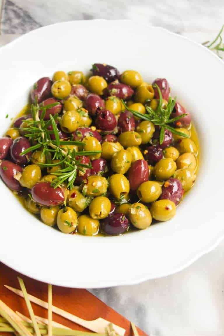 Close up of a white scalloped bowl of marinated olives, with skewers on the side.