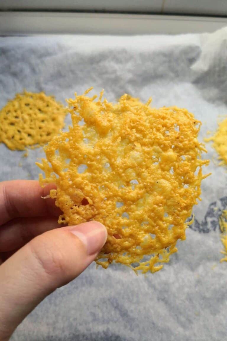 Hand holding up a parmesan crisp with more parmesan crisps on an oven tray in the background.