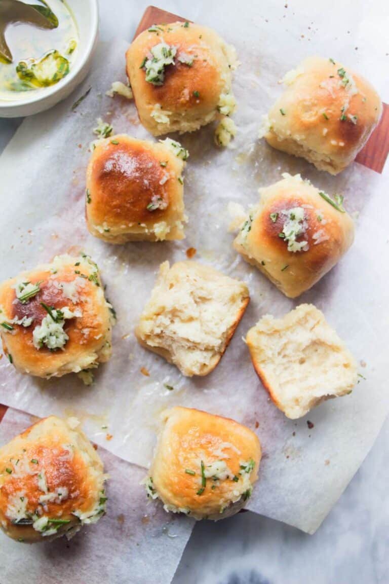 8 garlic butter topped rolled assembled on a baking paper lined wooden board with bowl of garlic butter in the background and one roll torn open.