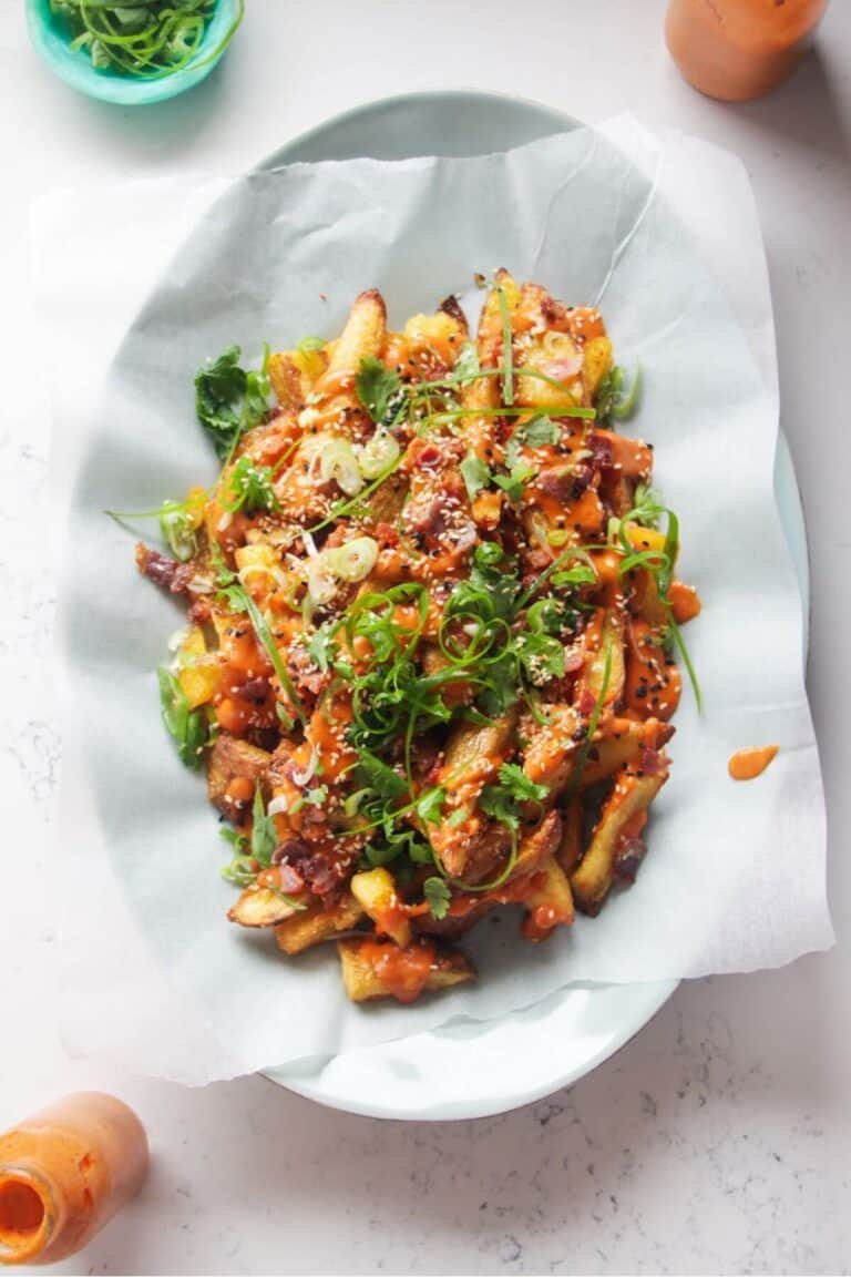 Loaded potato wedges on a large oval white plate with small jug of gochujang bechamel on the side.