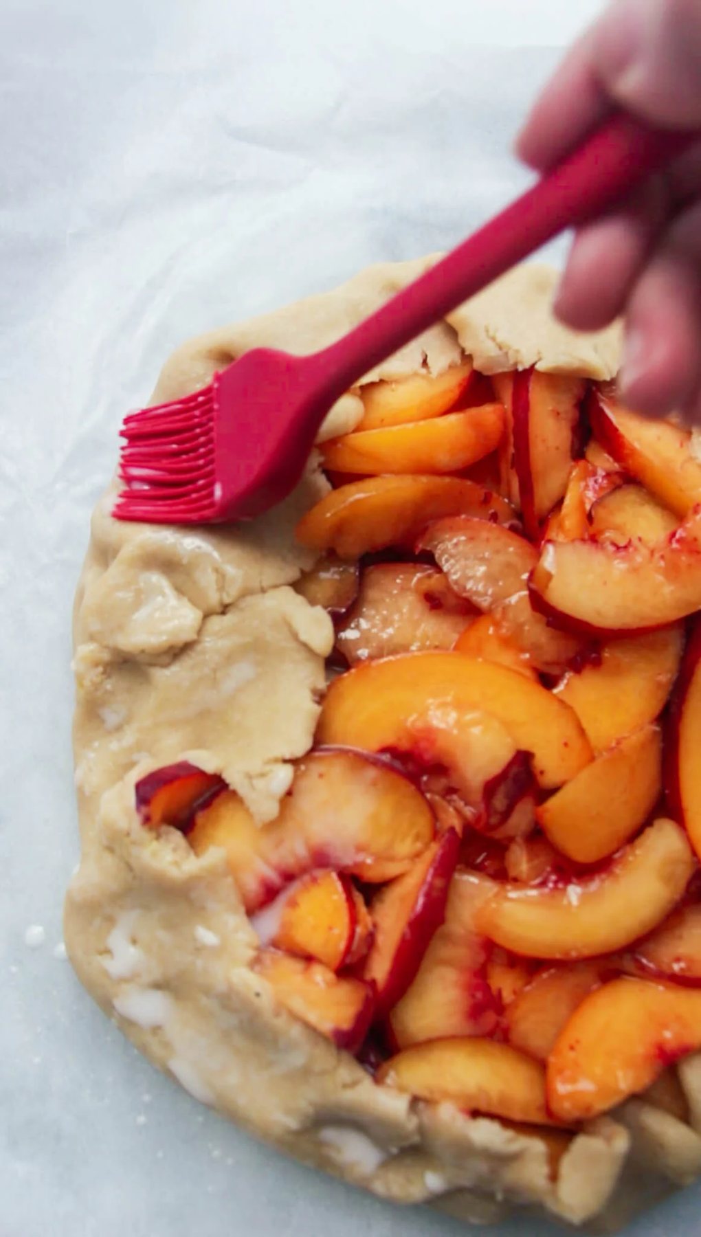 Red pastry brush brushing peach galette pastry with egg wash.